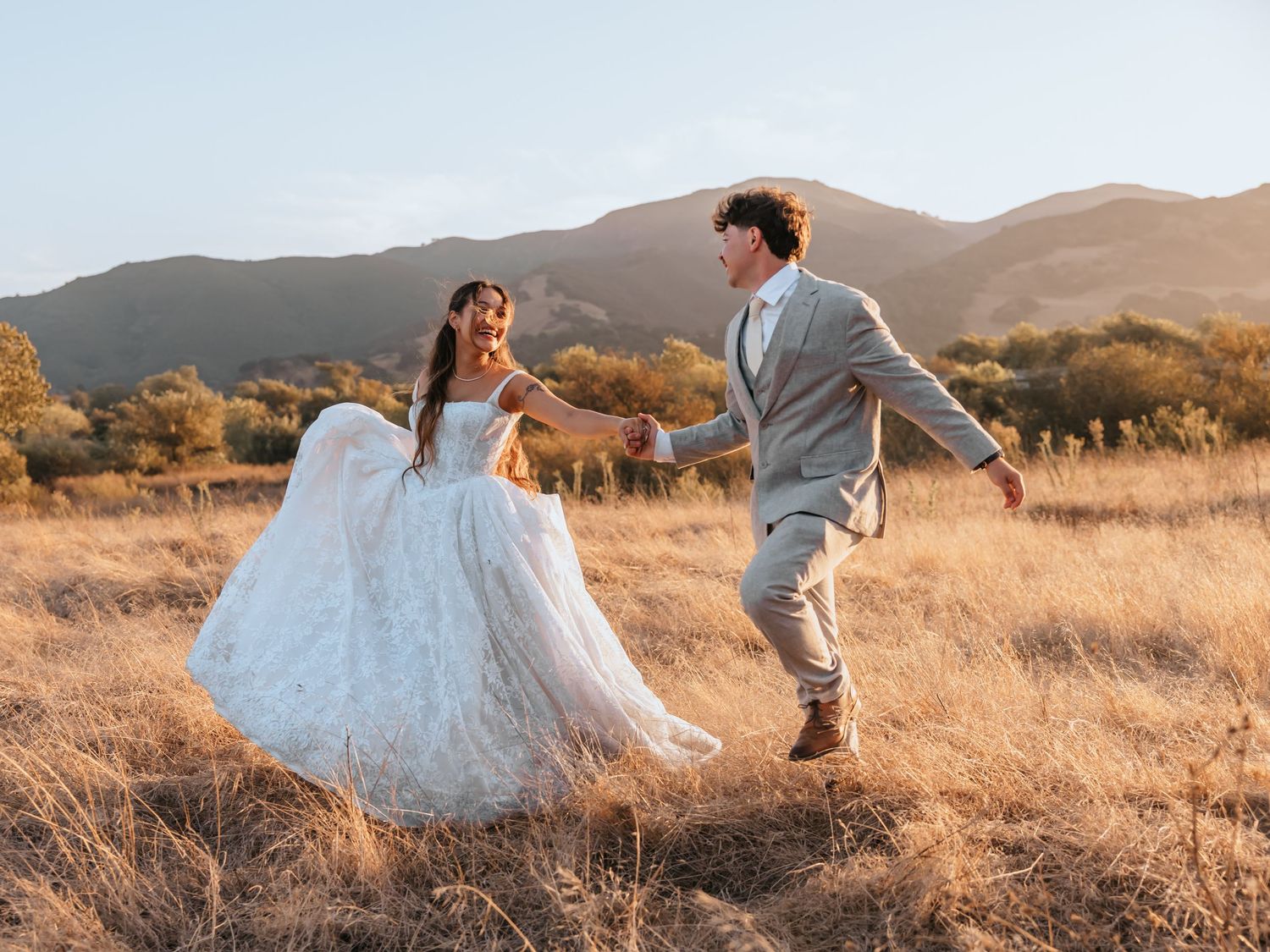 A bride and groom dance joyfully in a golden field with scenic mountains at sunset on their wedding day.