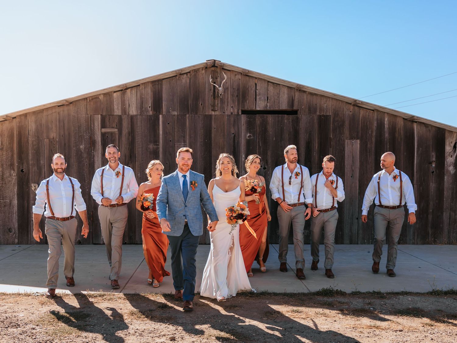 Wedding party dressed in rustic attire with suspenders and orange dresses pose in front of a rustic wooden barn exterior.