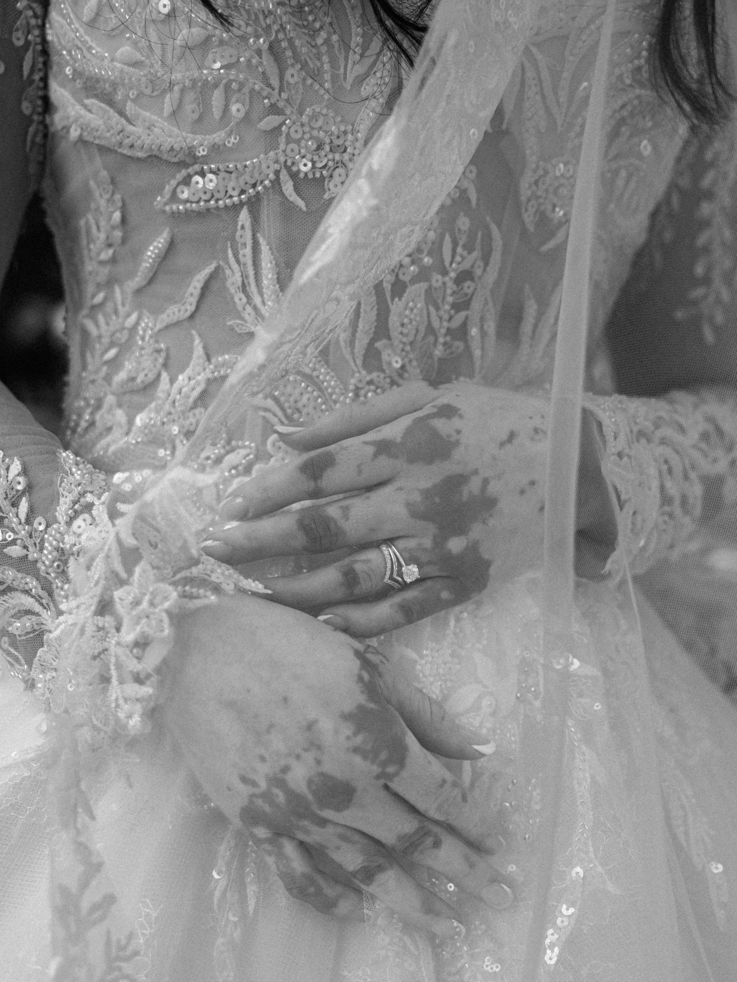Black and white close-up of bride's hands with wedding ring on lace embroidered gown with flowing veil.