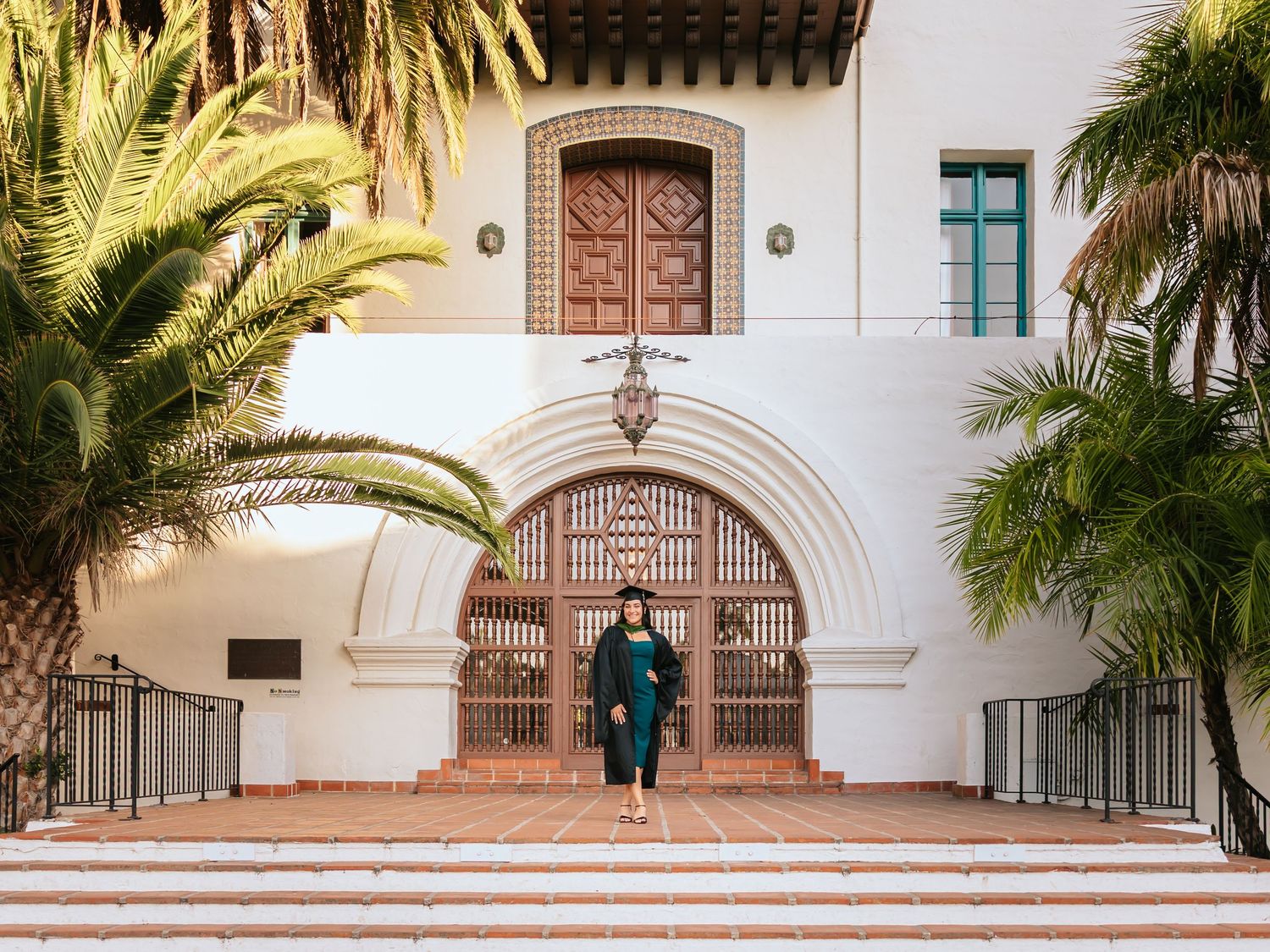 Woman in teal dress standing on steps of a Spanish Colonial Revival building with palm trees.