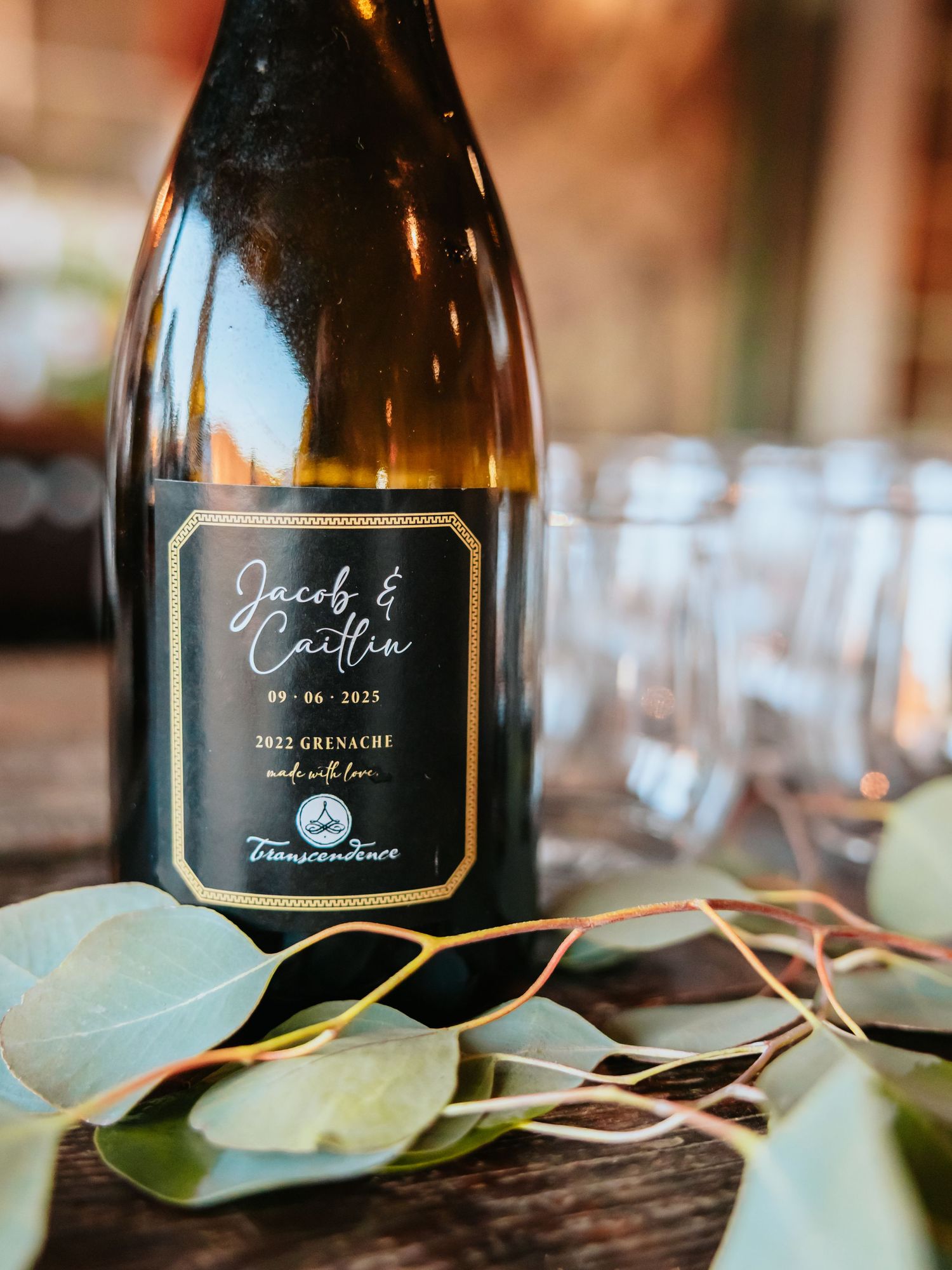 Custom labeled Jacob & Caitlin 2022 Grenache wine bottle with gold script, surrounded by eucalyptus leaves on a rustic table.