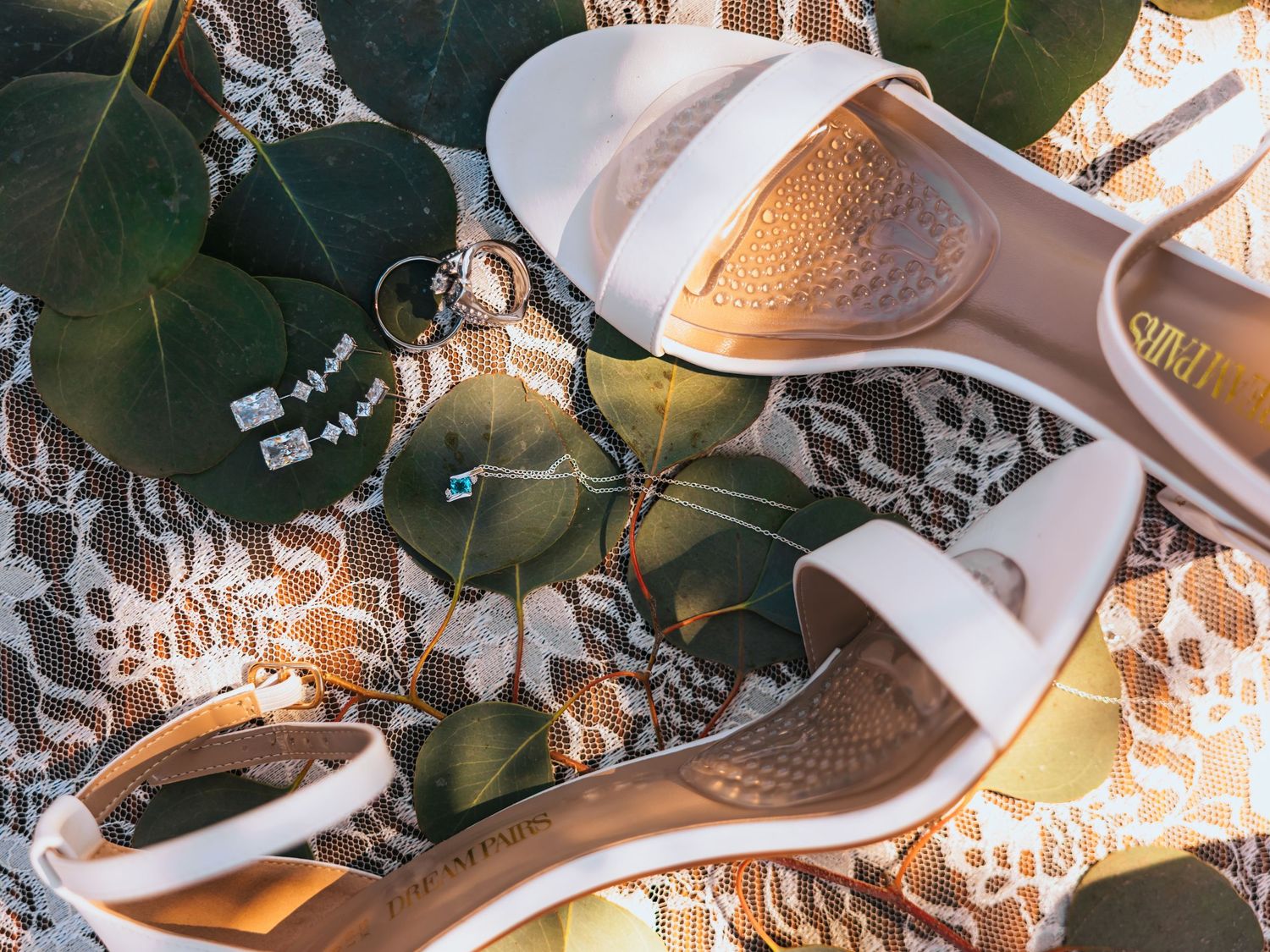 White bridal heeled sandals with gold insoles on lace fabric surrounded by green leaves and delicate jewelry.