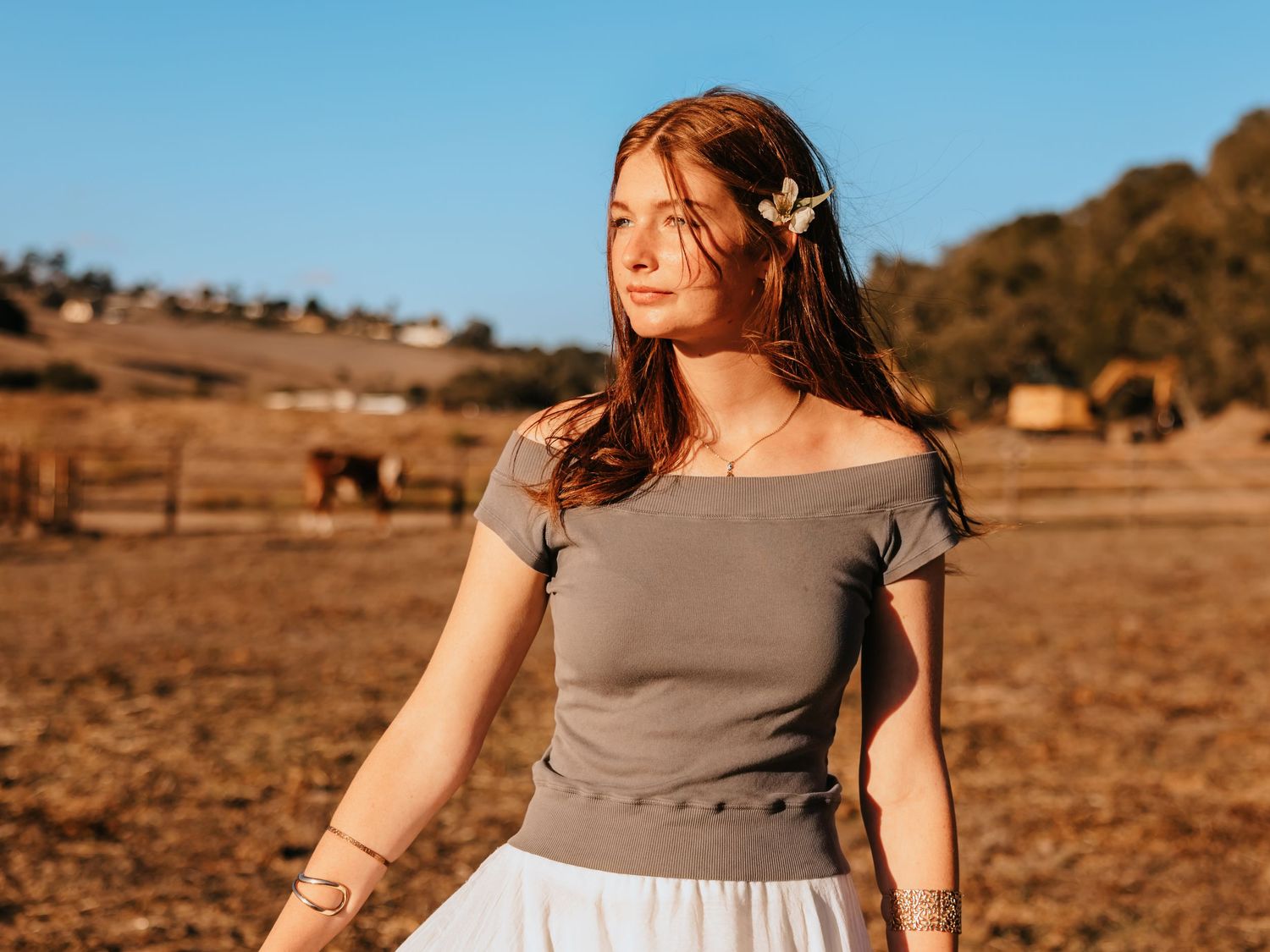 Young woman with red hair and flower accessory wearing gray off-shoulder top and white skirt in sunny farm field.