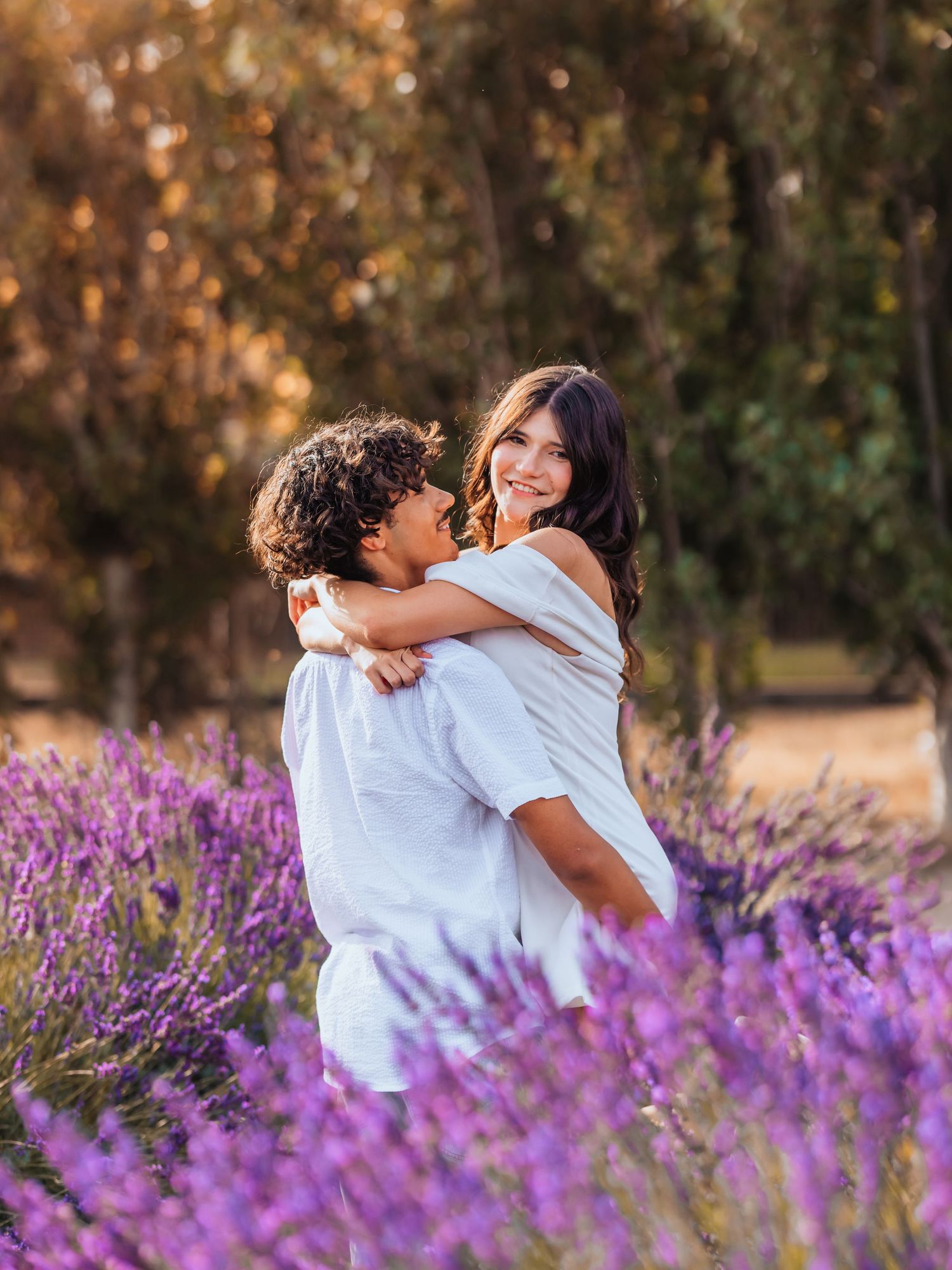 Couple embracing in a lavender field, woman smiling over mans shoulder, surrounded by purple blooms and autumn trees.