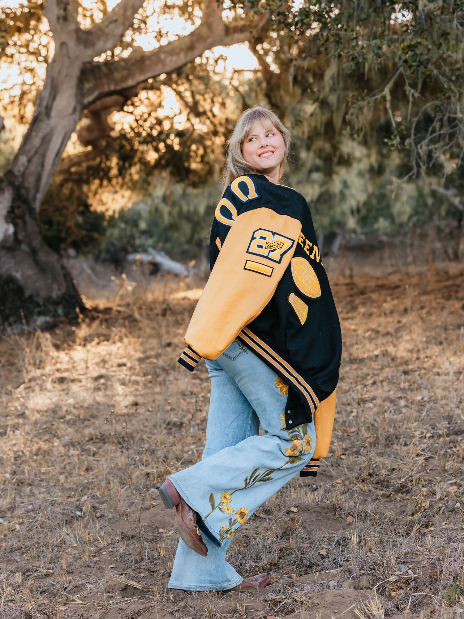 Woman wearing black and gold varsity jacket with light wash flare jeans outdoors in golden hour light.