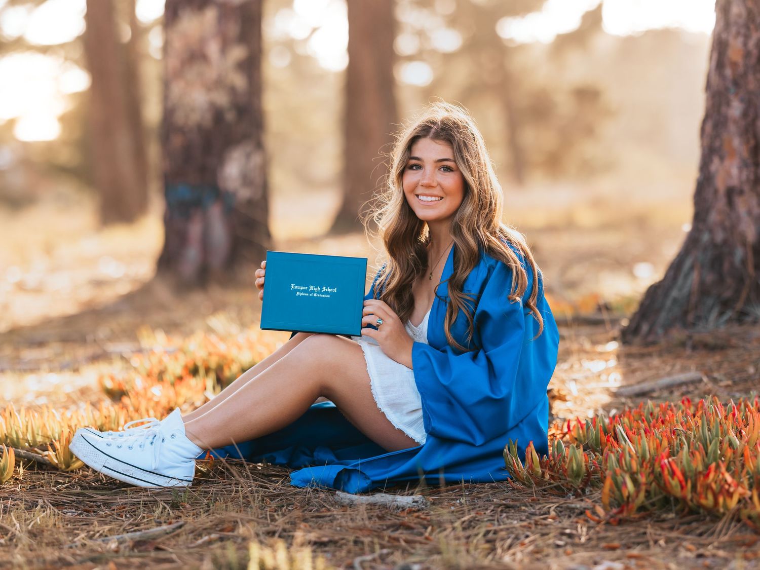 Someone in a bright blue dress sits on autumn ground with a book while wearing white sneakers in a sunlit forest setting.
