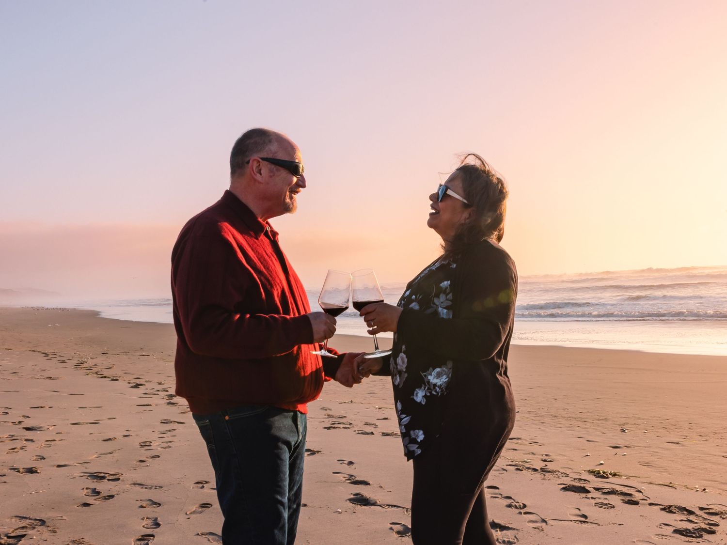 Two people sharing a conversation on a beach during sunset with pink-orange skies and ocean waves in the background.