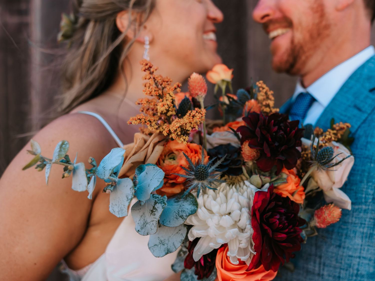 Smiling bride and groom holding a vibrant fall wedding bouquet with burgundy, orange, and white blooms and eucalyptus.