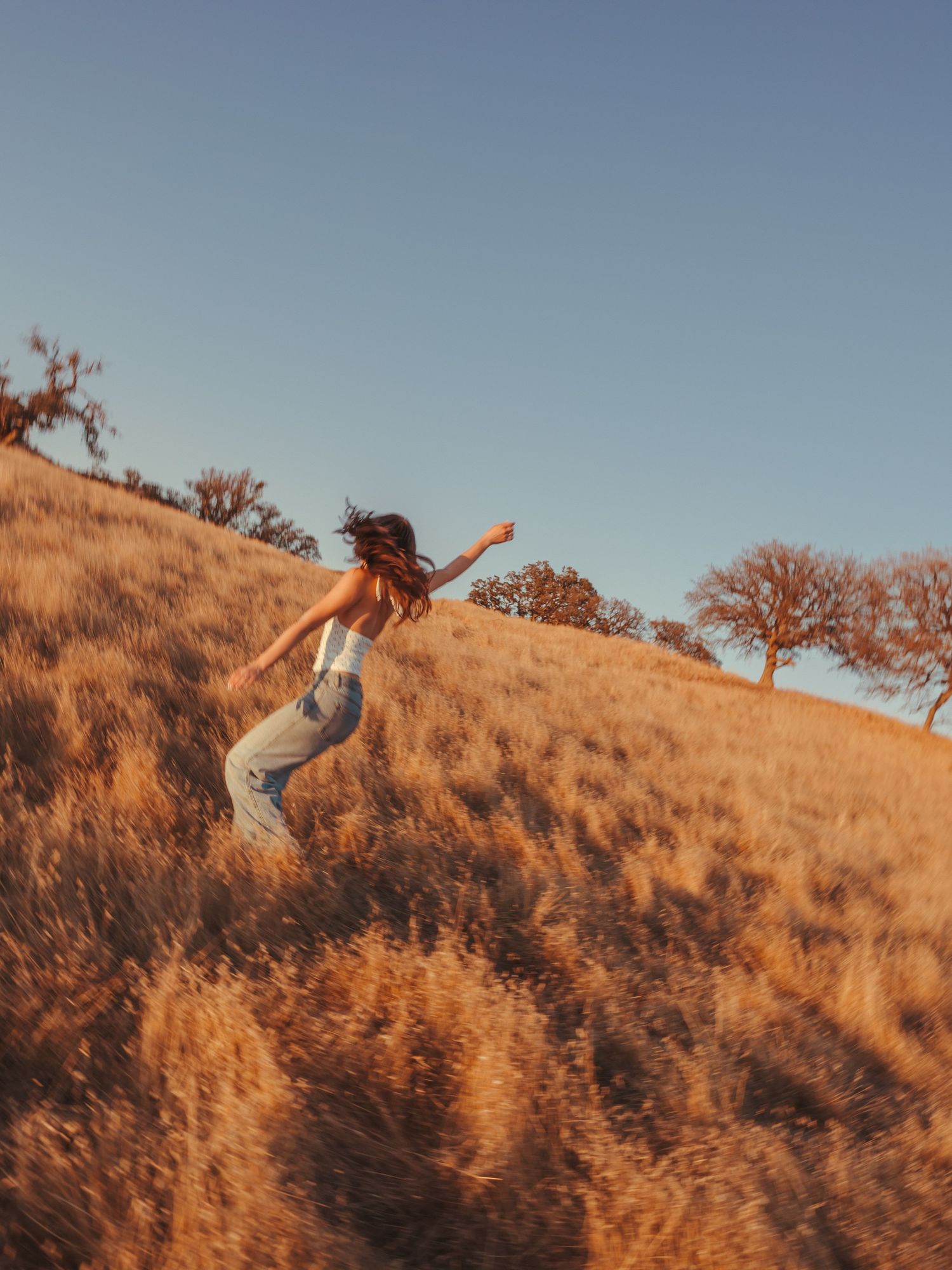 A woman in a white top and jeans runs through golden dry grass on a sunlit hillside with trees in the background.