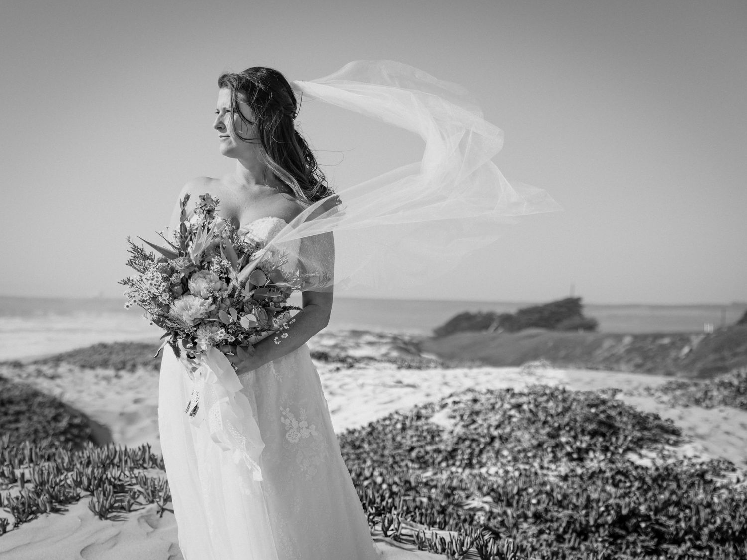 Black and white scenic coastal wedding photo showing figure in flowing dress with bouquet as veil catches the ocean breeze.