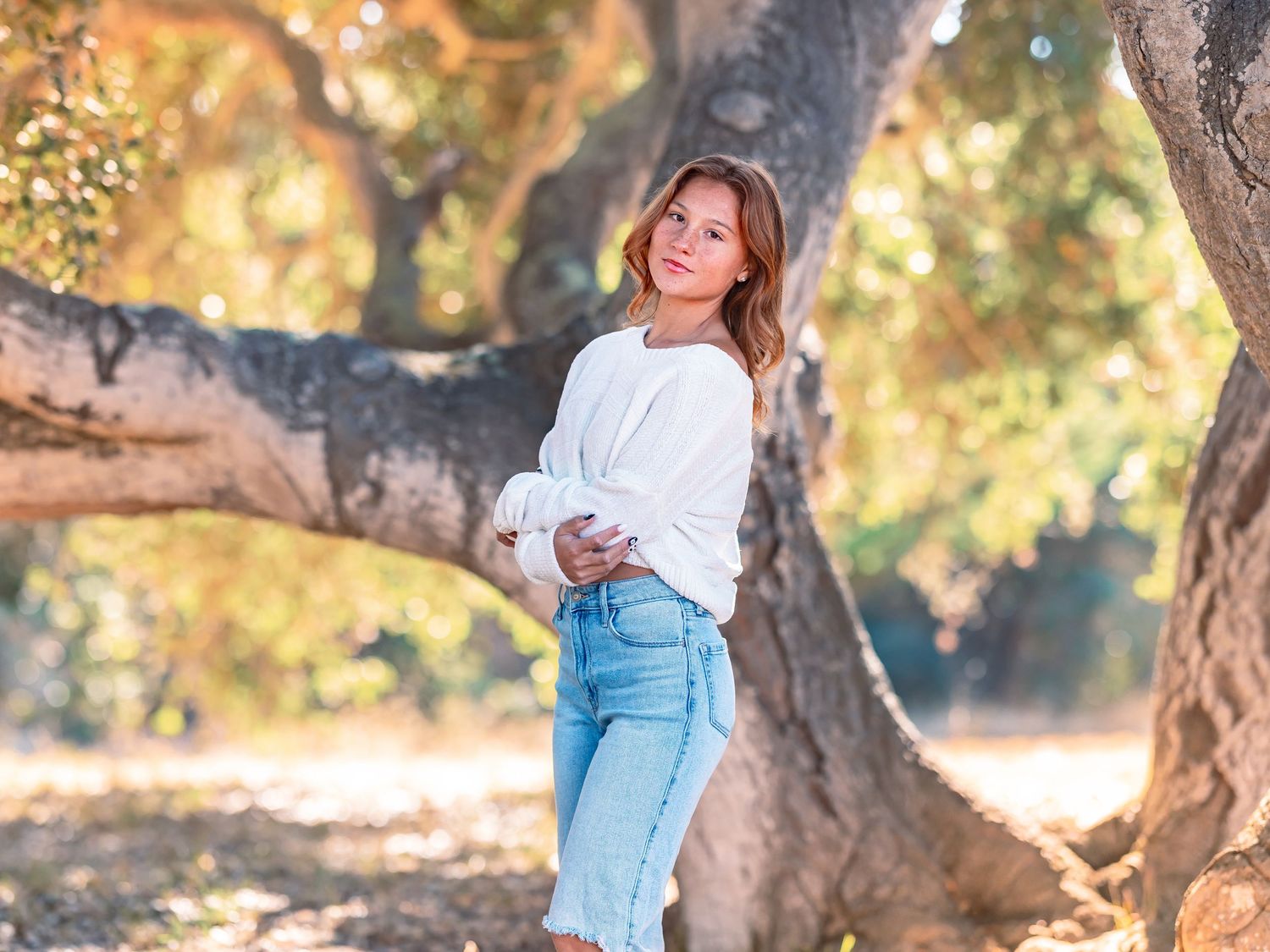 Young woman in white sweater and jeans posing by a large tree in a sunlit outdoor setting.
