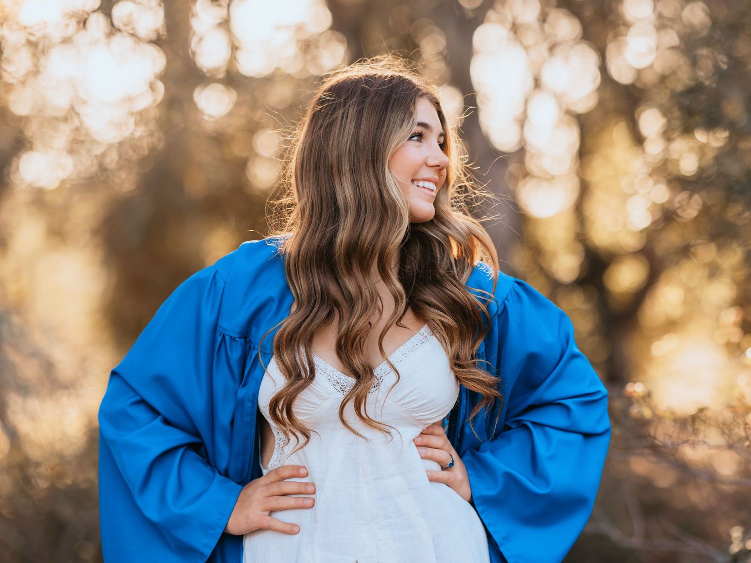 Joyful person in a bright blue jacket and white top smiles during golden hour with bokeh-lit trees in background.
