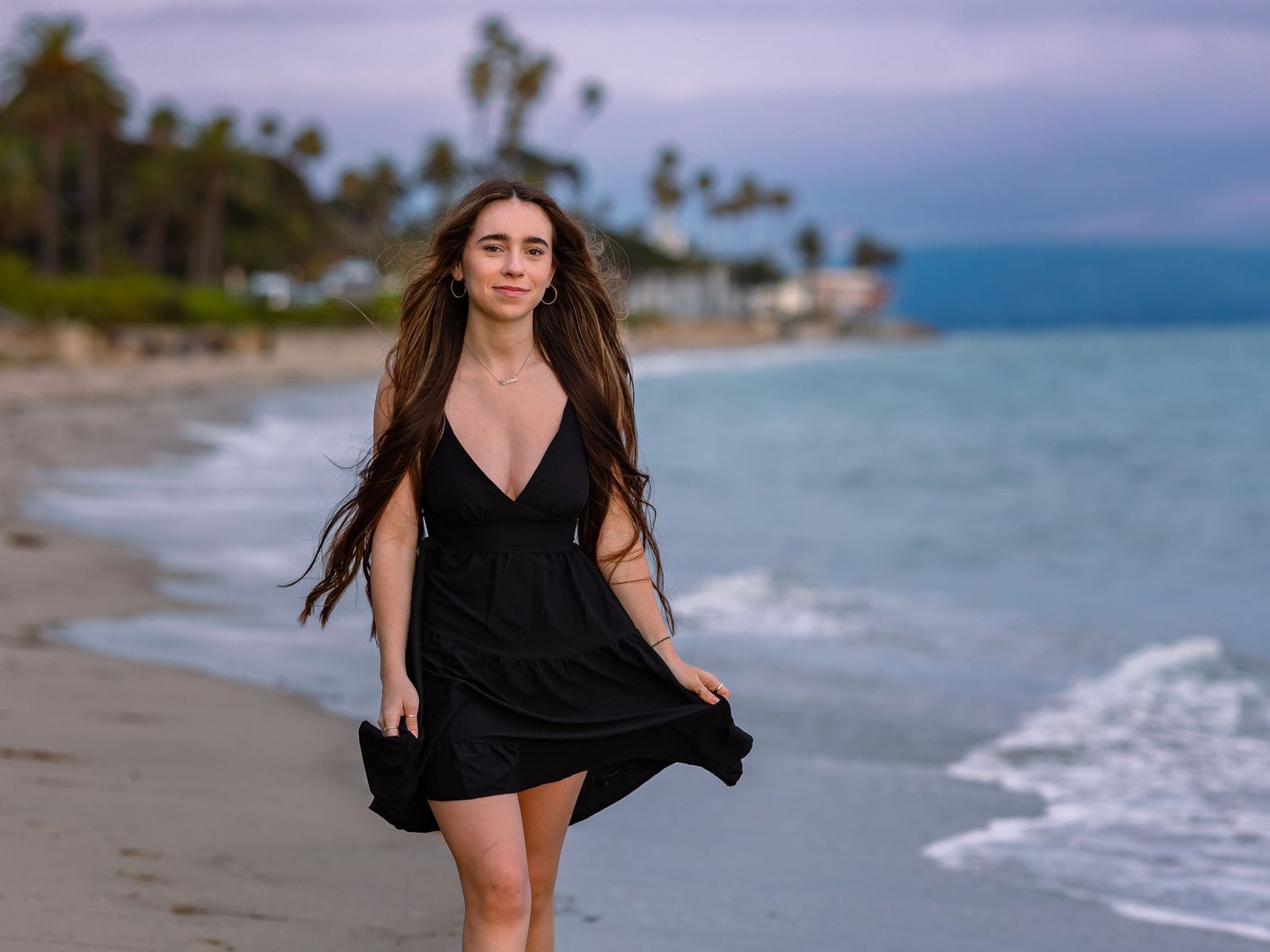 Young woman in black dress walking on tropical beach with palm trees and turquoise ocean waves in background.
