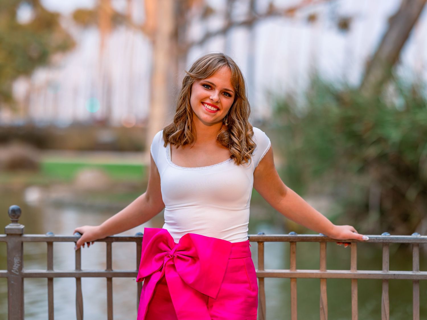 Young woman in white top and pink bow skirt posing by iron railing near a pond outdoors.