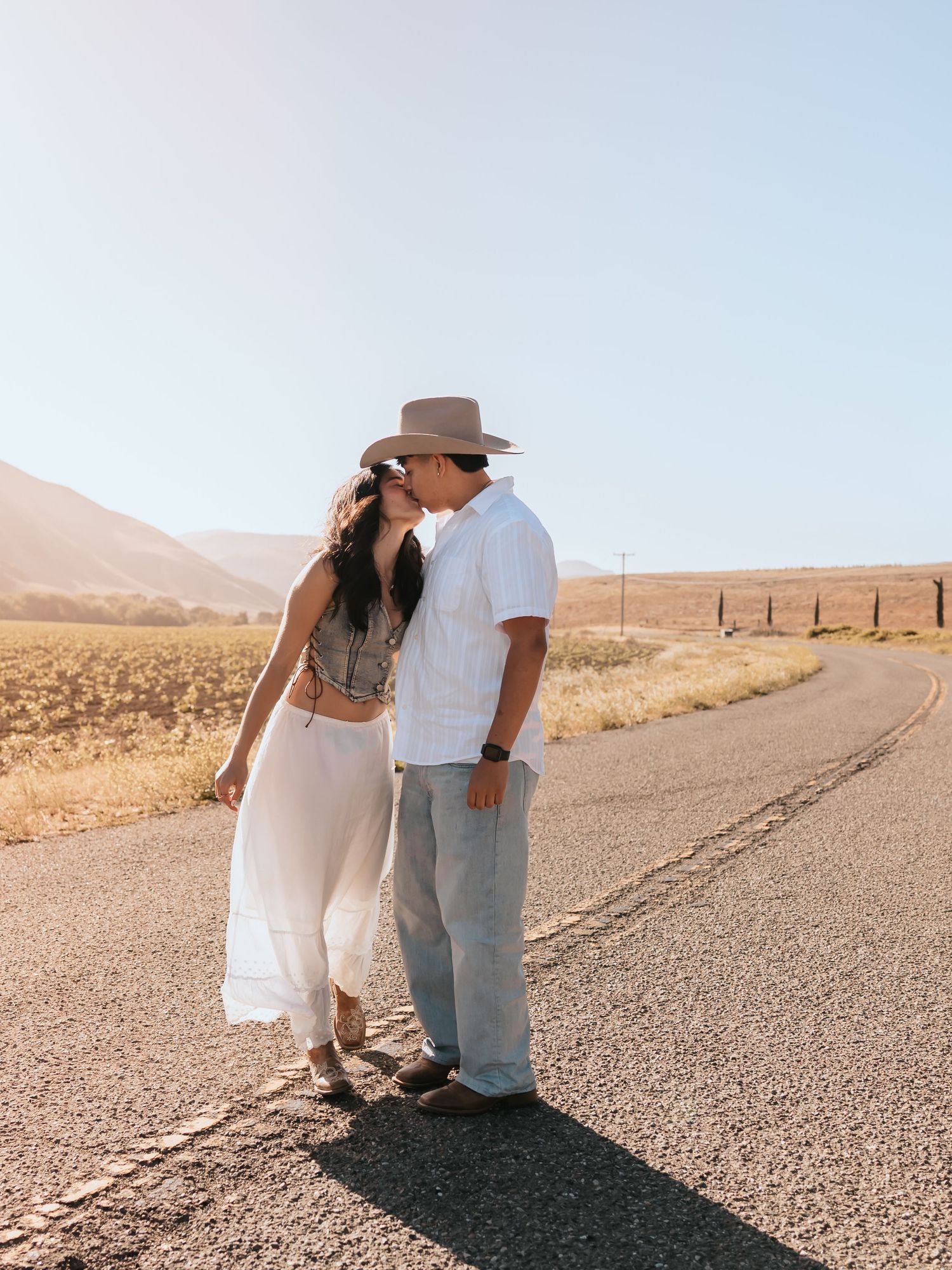 Couple sharing a romantic kiss on a desert road, woman in white skirt and man in hat, with golden hills backdrop.