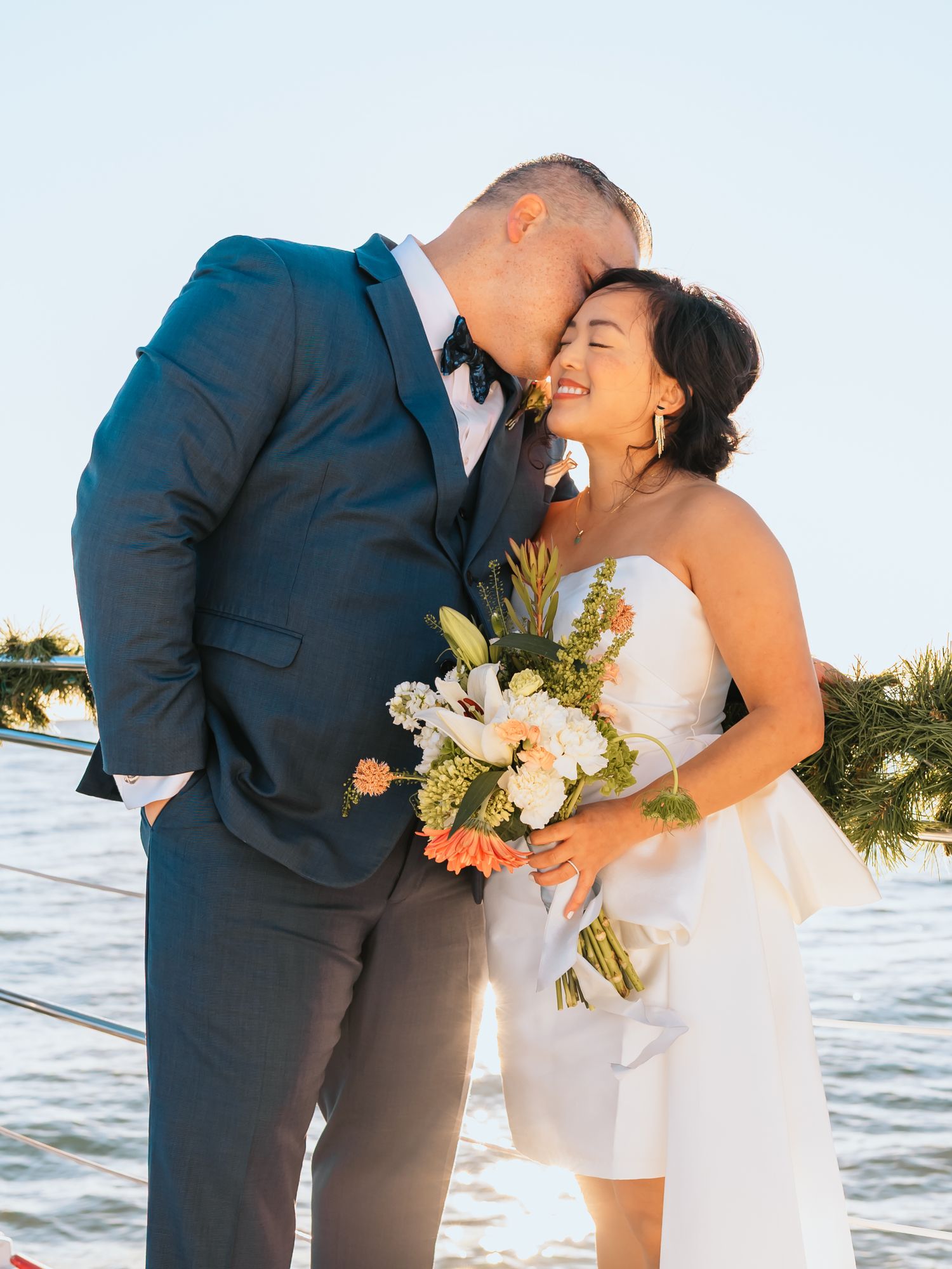 Bride and groom share a romantic moment on a waterfront dock, bride holding a white and coral bouquet in a strapless dress.