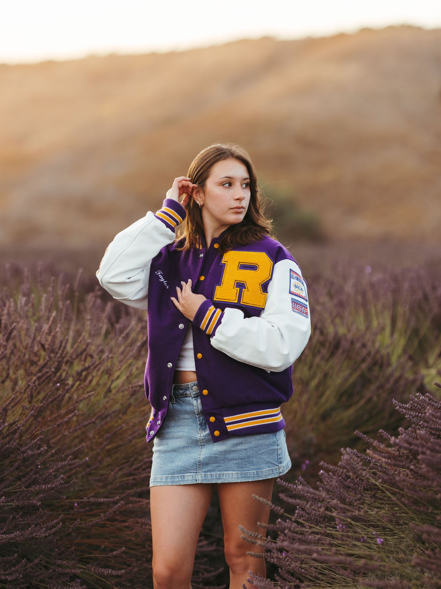 Young woman in purple varsity jacket and denim skirt posing in lavender field at golden hour.