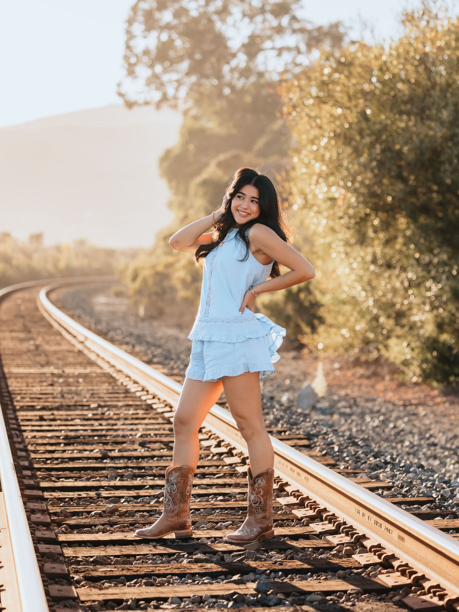 Young woman in light blue dress and cowboy boots posing on railroad tracks with golden hour sunlight.