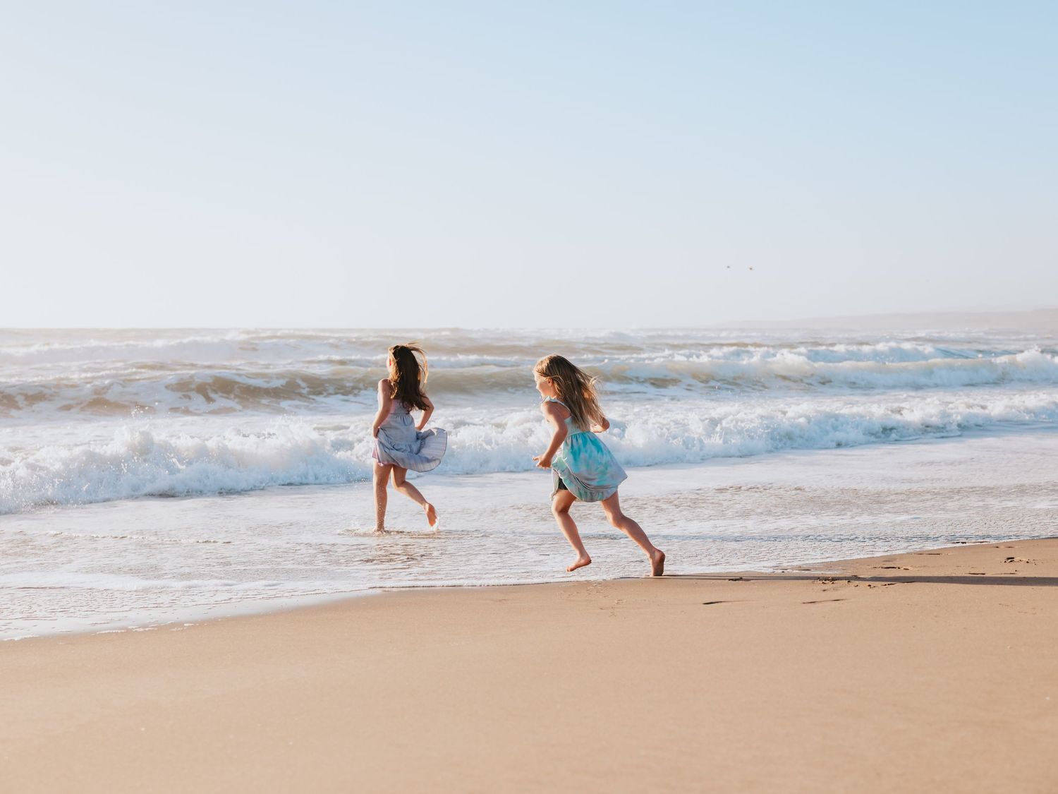 Two people run joyfully along a sandy beach with crashing ocean waves in the background on a sunny day.