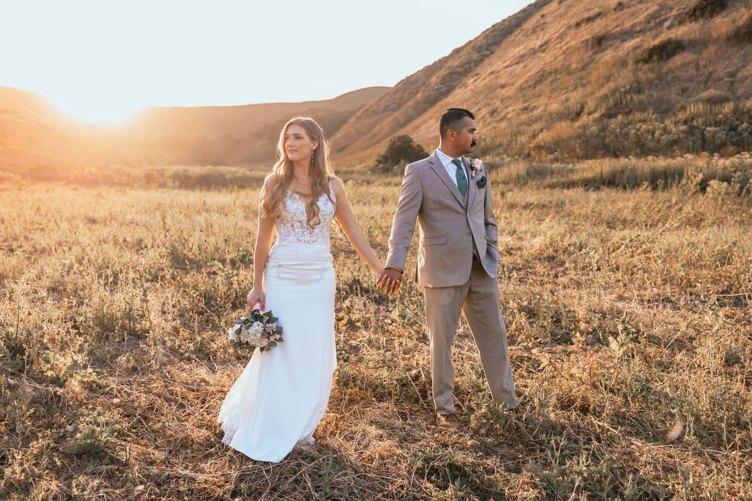 Bride and groom holding hands walking through golden hillside field at sunset.