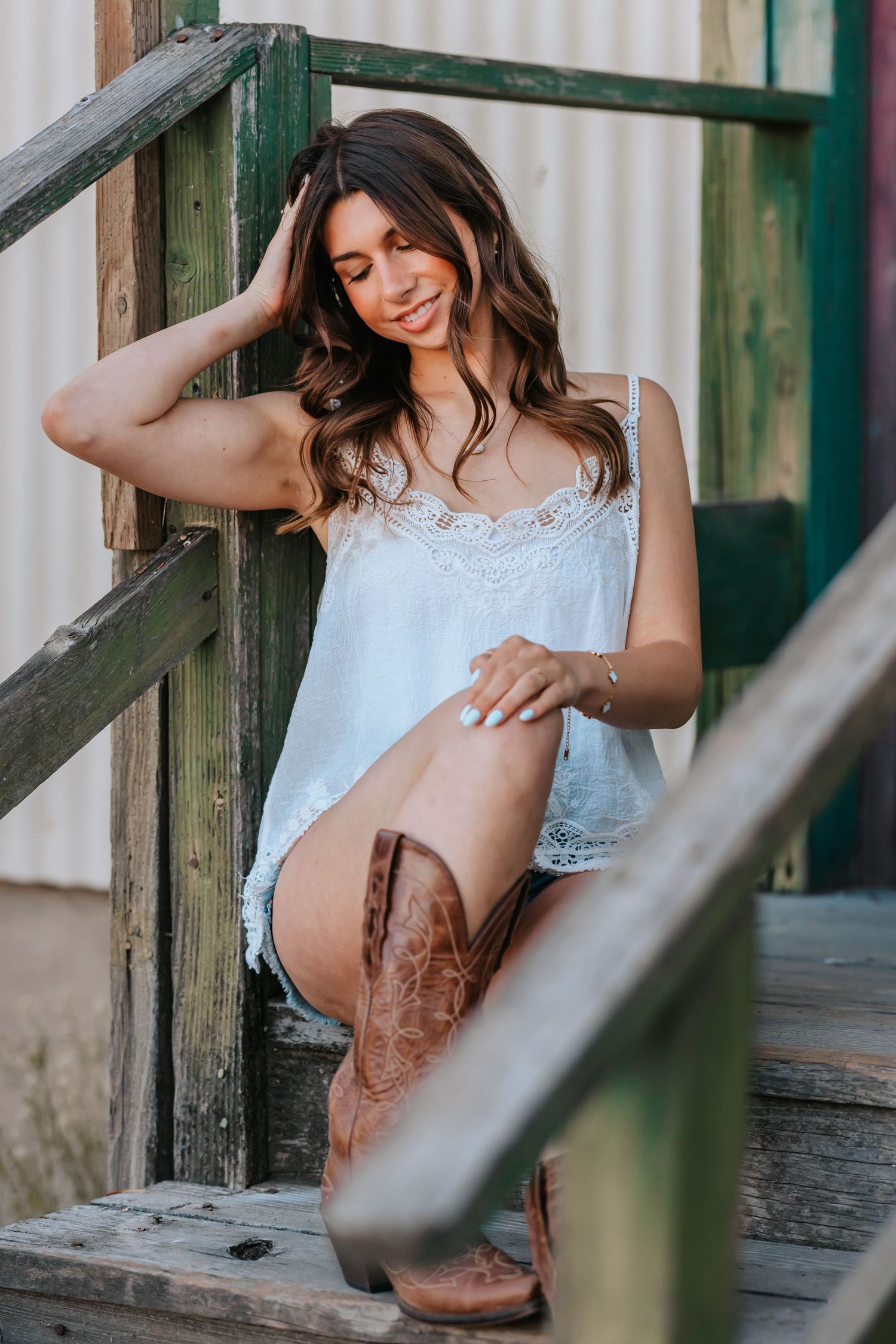 Young woman in white lace top, denim shorts, and cowboy boots sitting on rustic wooden steps smiling.