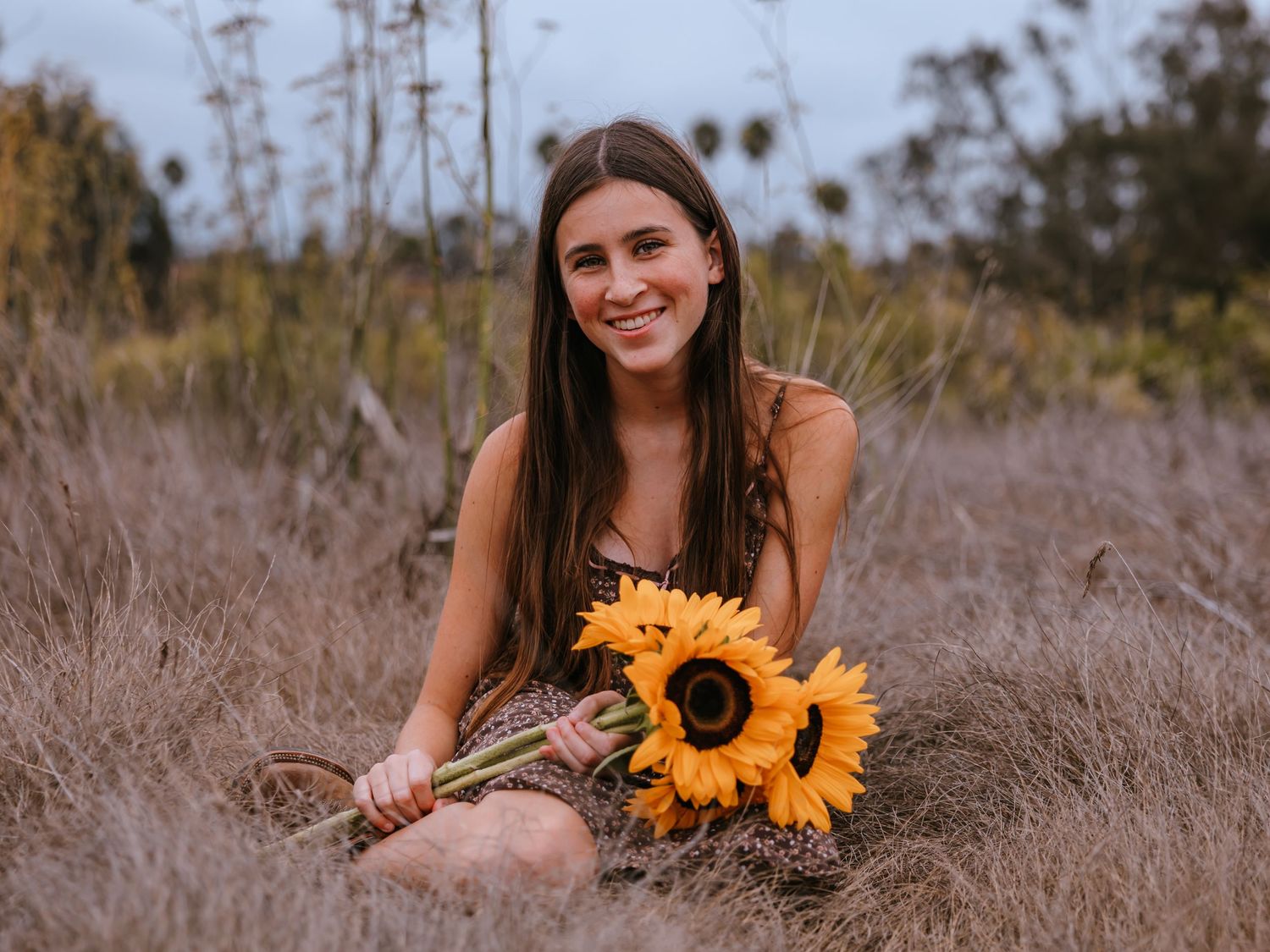 Young woman sitting in a golden field holding vibrant sunflowers, smiling outdoors in natural light.