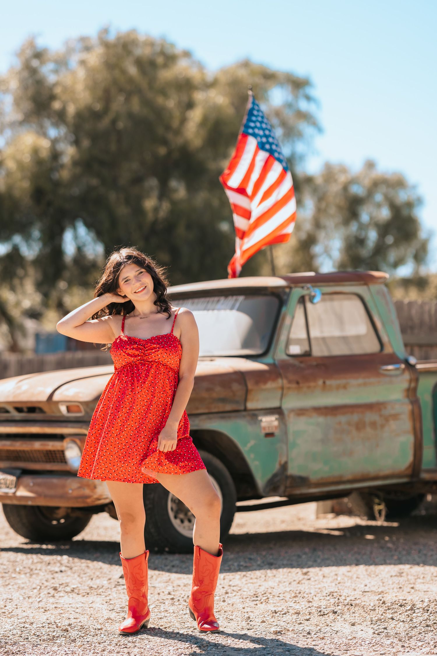 A young woman in a red dress and red cowboy boots stands before a rusty truck and American flag.
