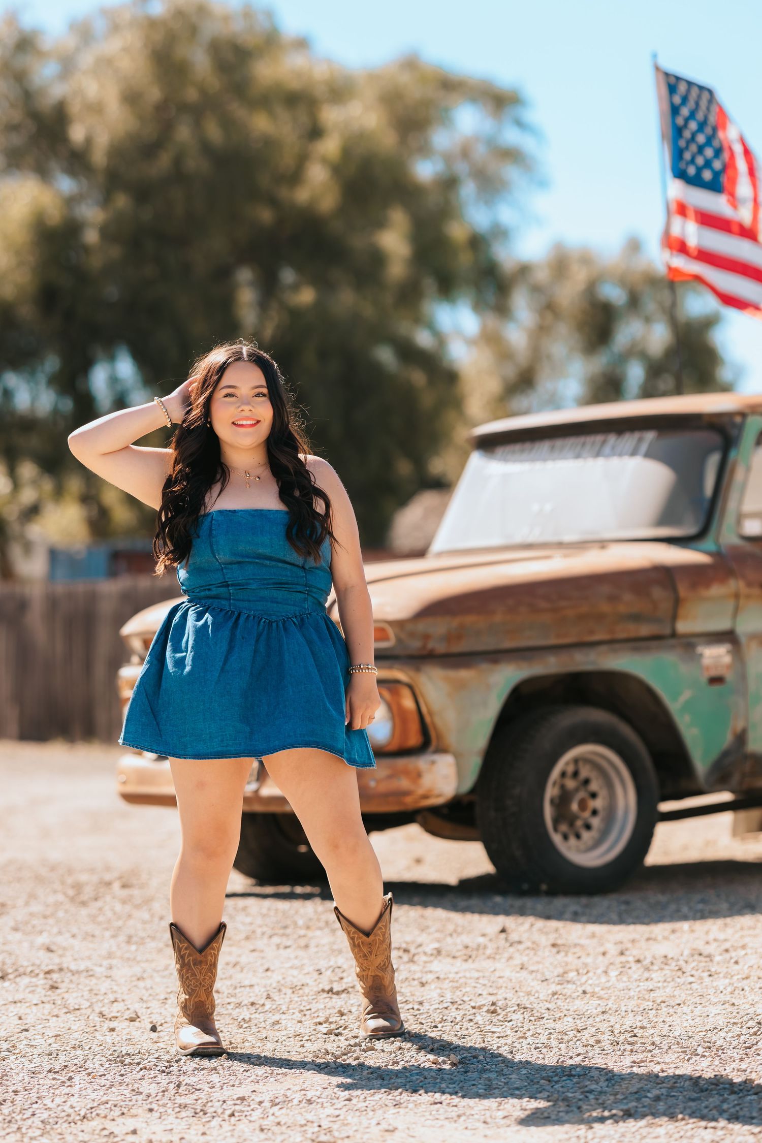A smiling young woman in a blue dress and cowboy boots poses near a vintage rusty truck.