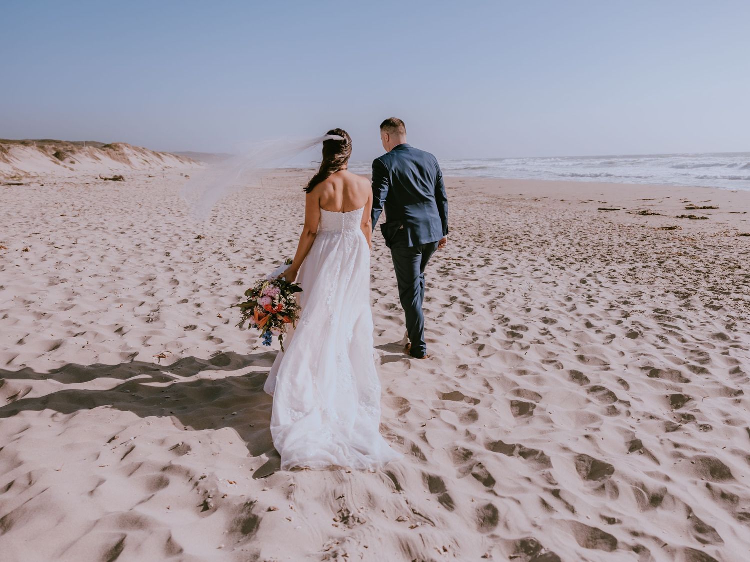 A couple walking on a sandy beach with rolling dunes, the figure in white holding a vibrant floral bouquet.