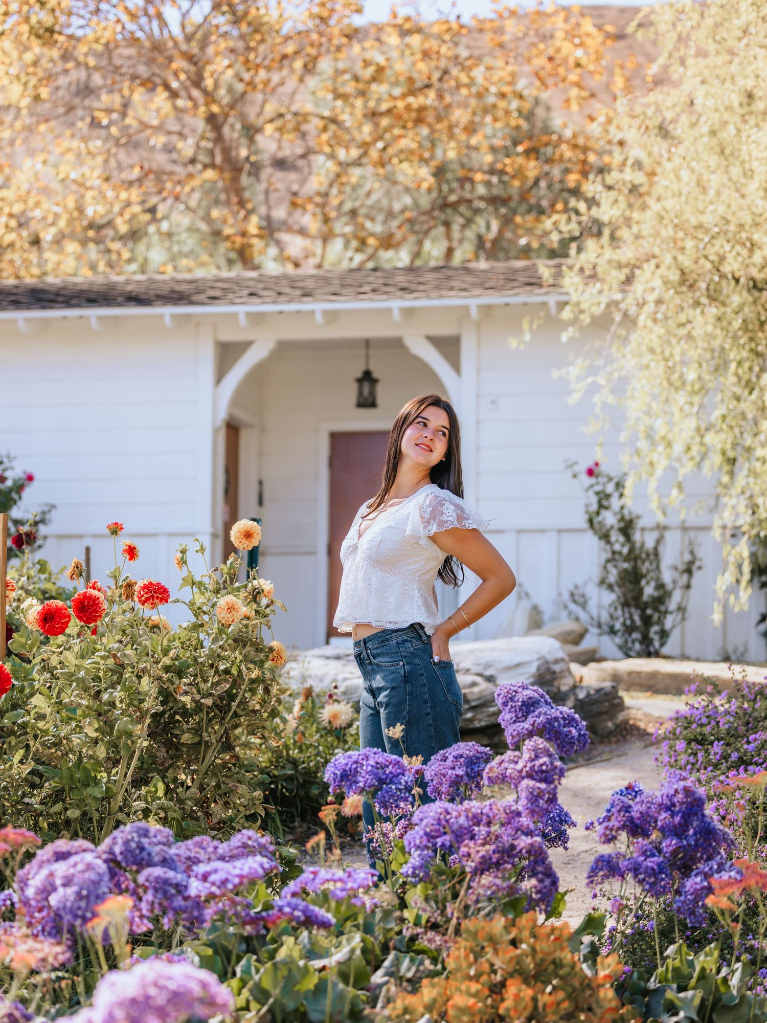 Woman in white top and jeans posing among colorful purple and red flowers in a garden near a white cottage.