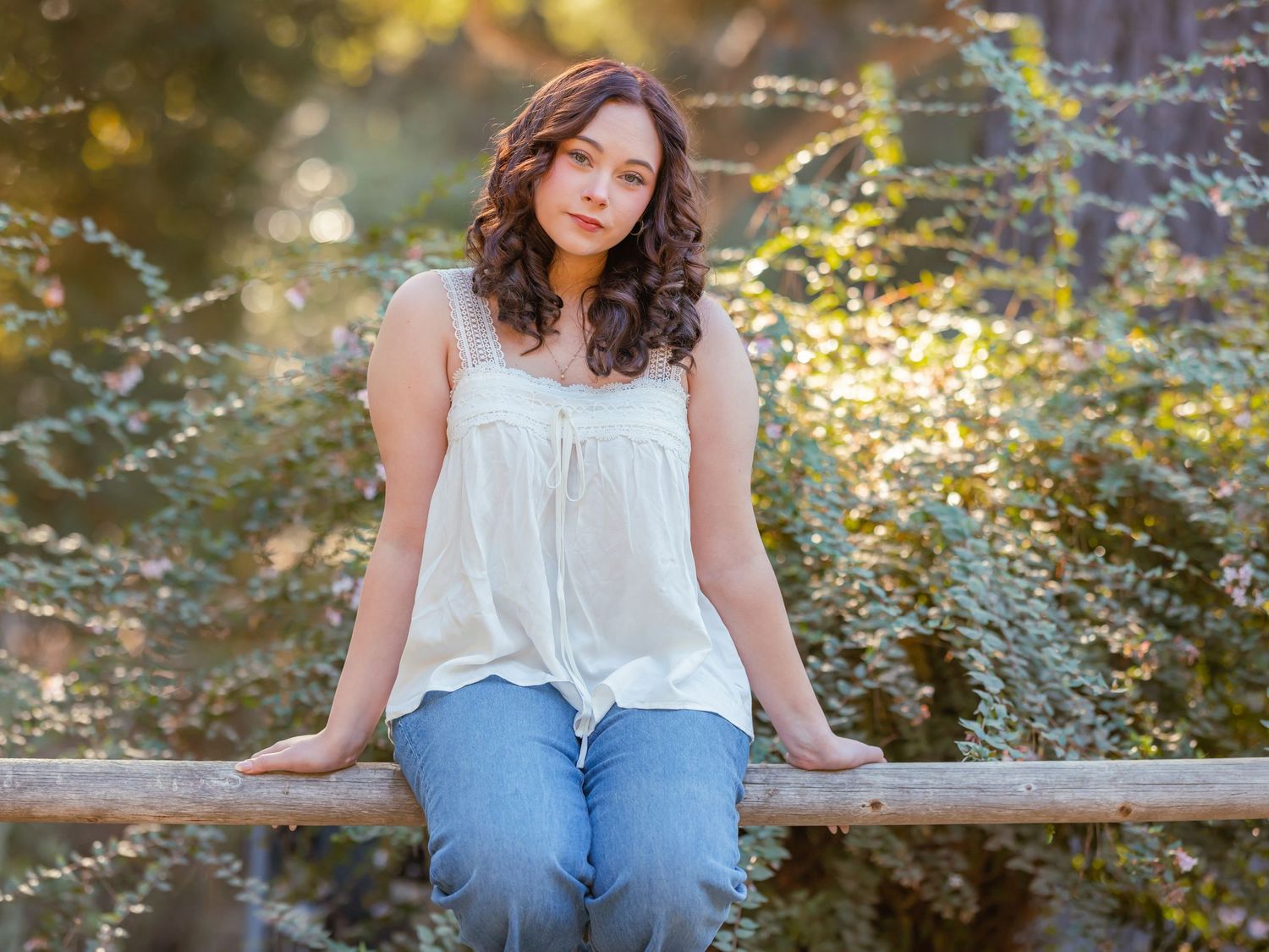 Young woman in white top and jeans sitting on wooden fence surrounded by lush green foliage outdoors.
