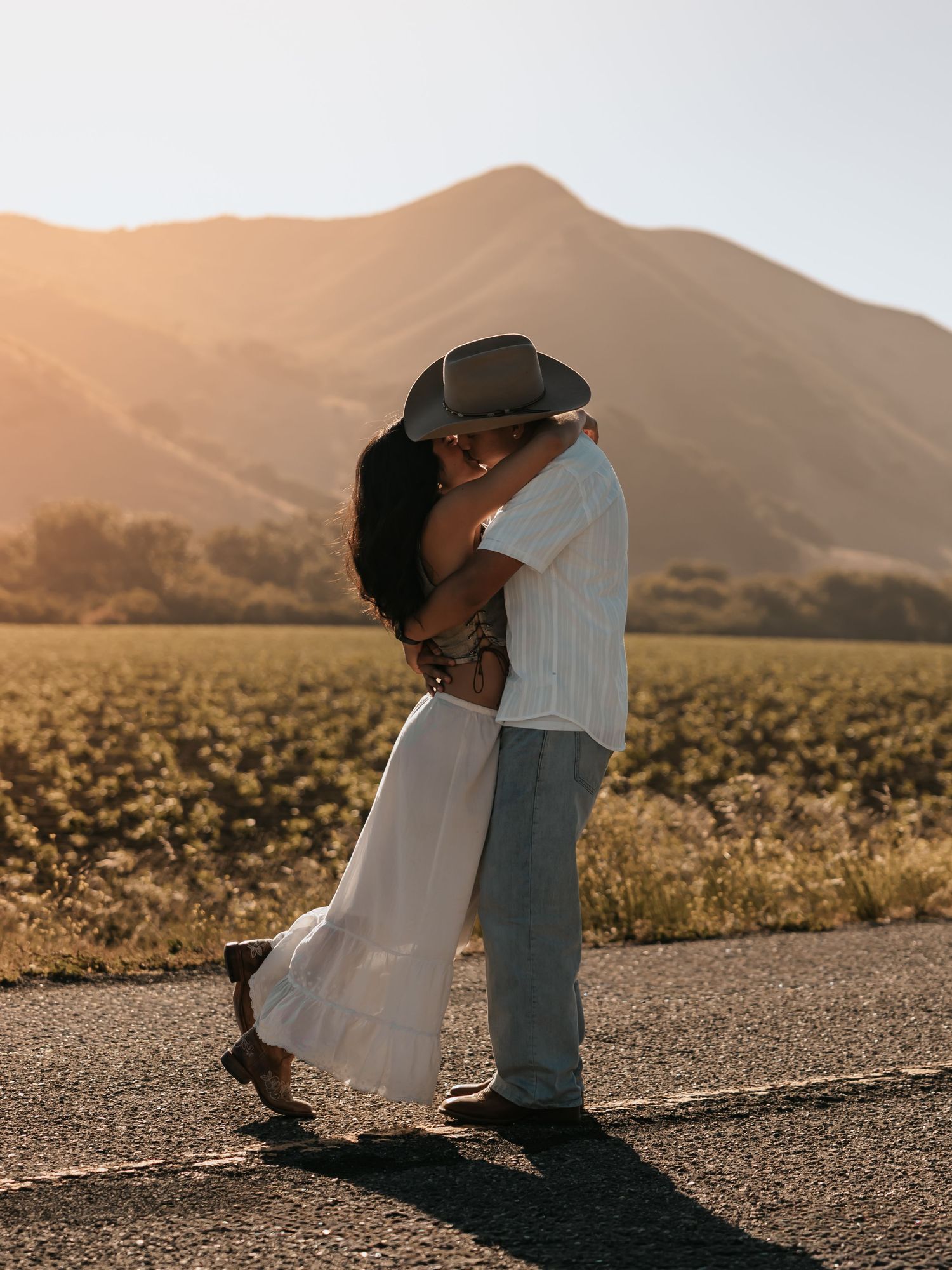 Couple sharing a romantic kiss on a road with golden mountain sunset backdrop, woman in white dress, man in cowboy hat.