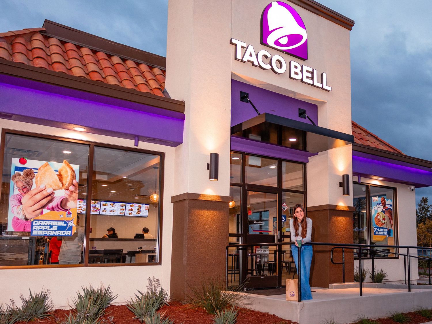 Taco Bell restaurant exterior at dusk with purple accent lighting, illuminated signage, and a woman standing at the entrance.