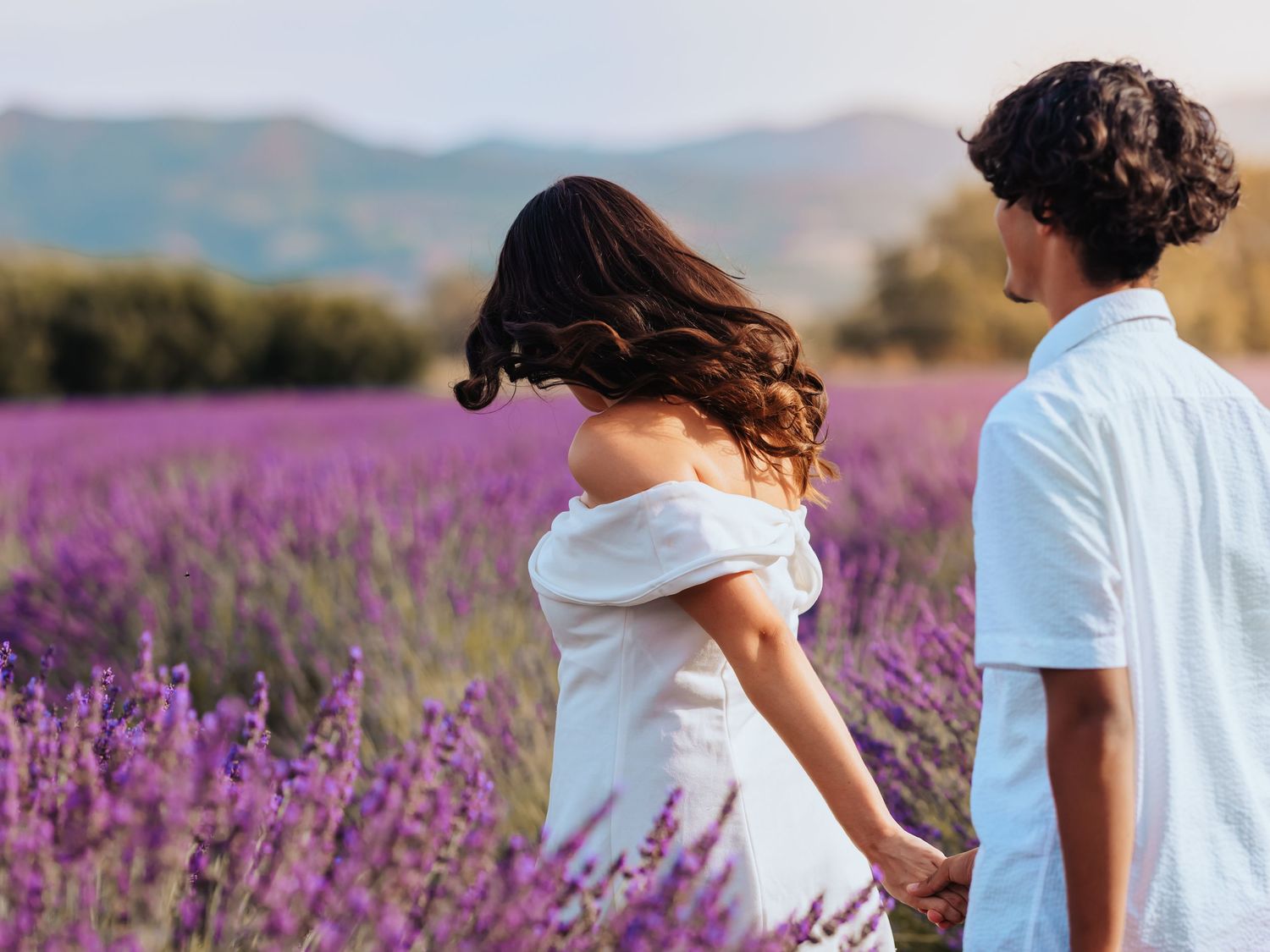 A couple walks hand in hand through a vibrant purple lavender field with rolling hills in the background.