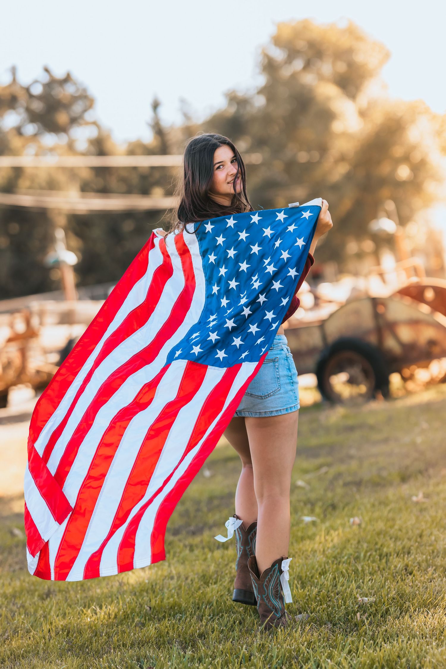 Young woman wrapped in large American flag wearing denim shorts and cowboy boots outdoors.