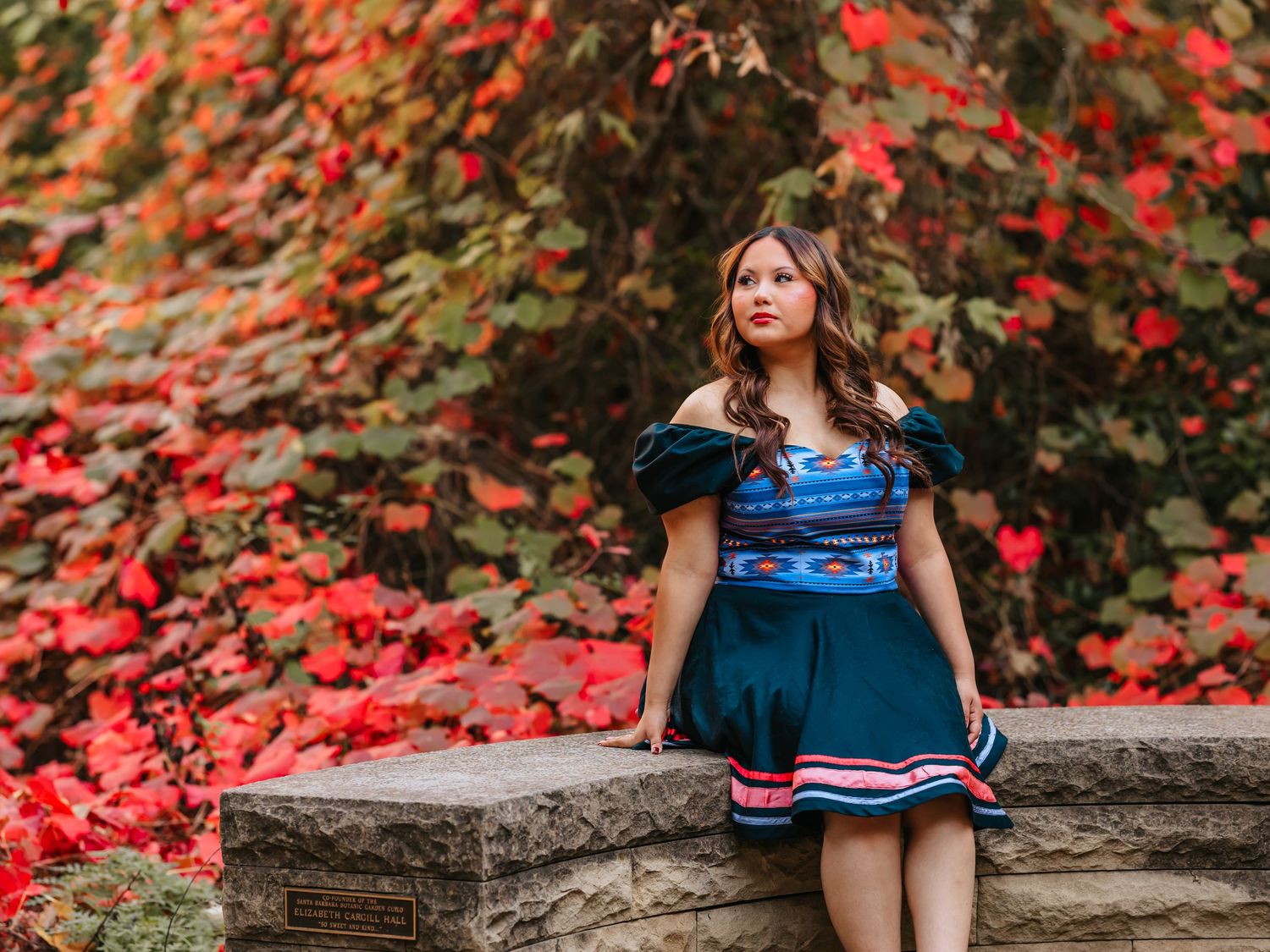 Young woman in blue patterned off-shoulder dress poses on stone wall with vibrant red autumn foliage backdrop.