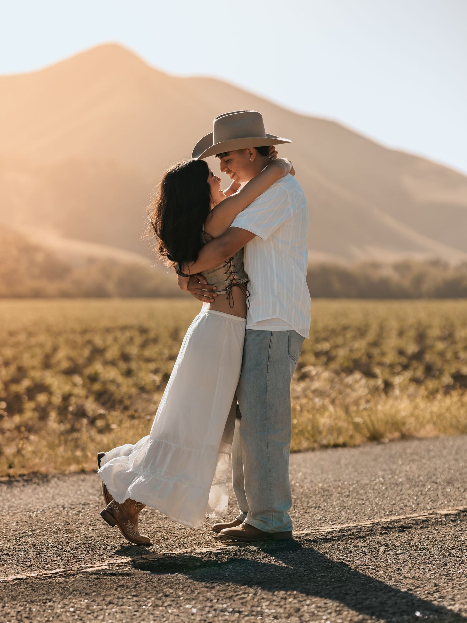 Couple sharing a romantic kiss on a sun-drenched road with mountain backdrop, woman in white dress, man in cowboy hat.