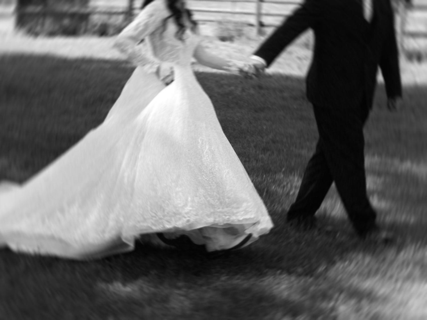 Black and white photo of a bride in a flowing white gown holding hands with a groom in a dark suit outdoors.