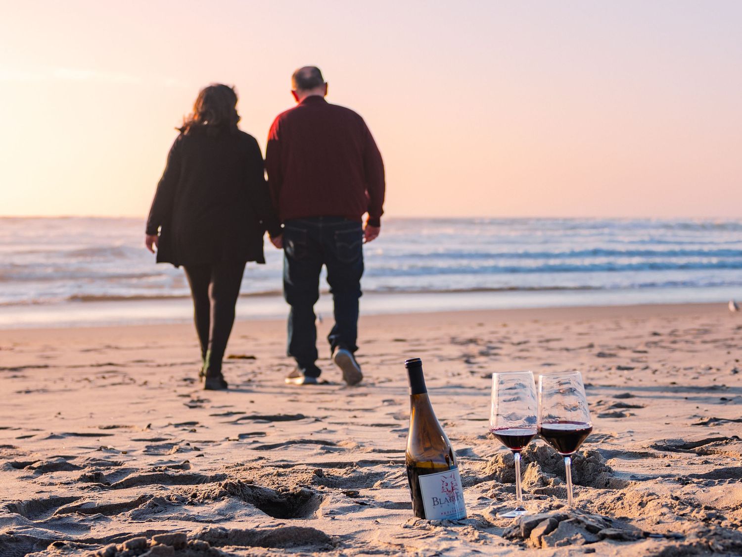 A bottle of wine and glasses on the beach at sunset with a couple walking away along the shoreline.