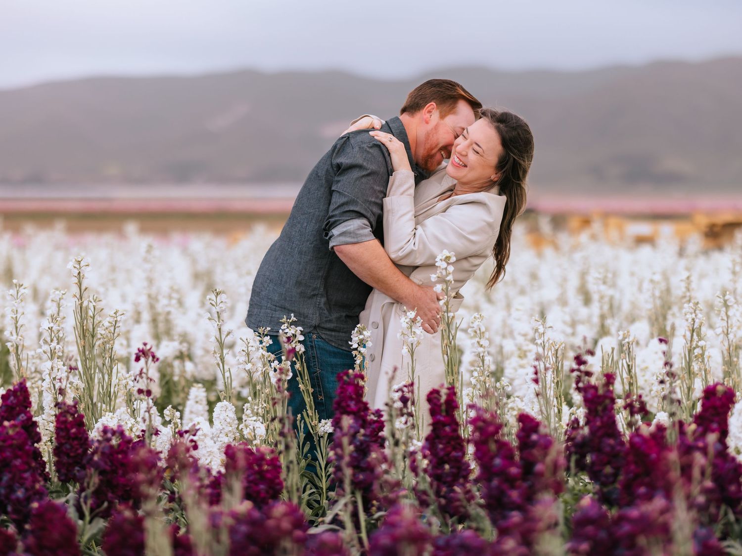 A couple embraces in a field of white and purple wildflowers with mountains in the distance at sunset.