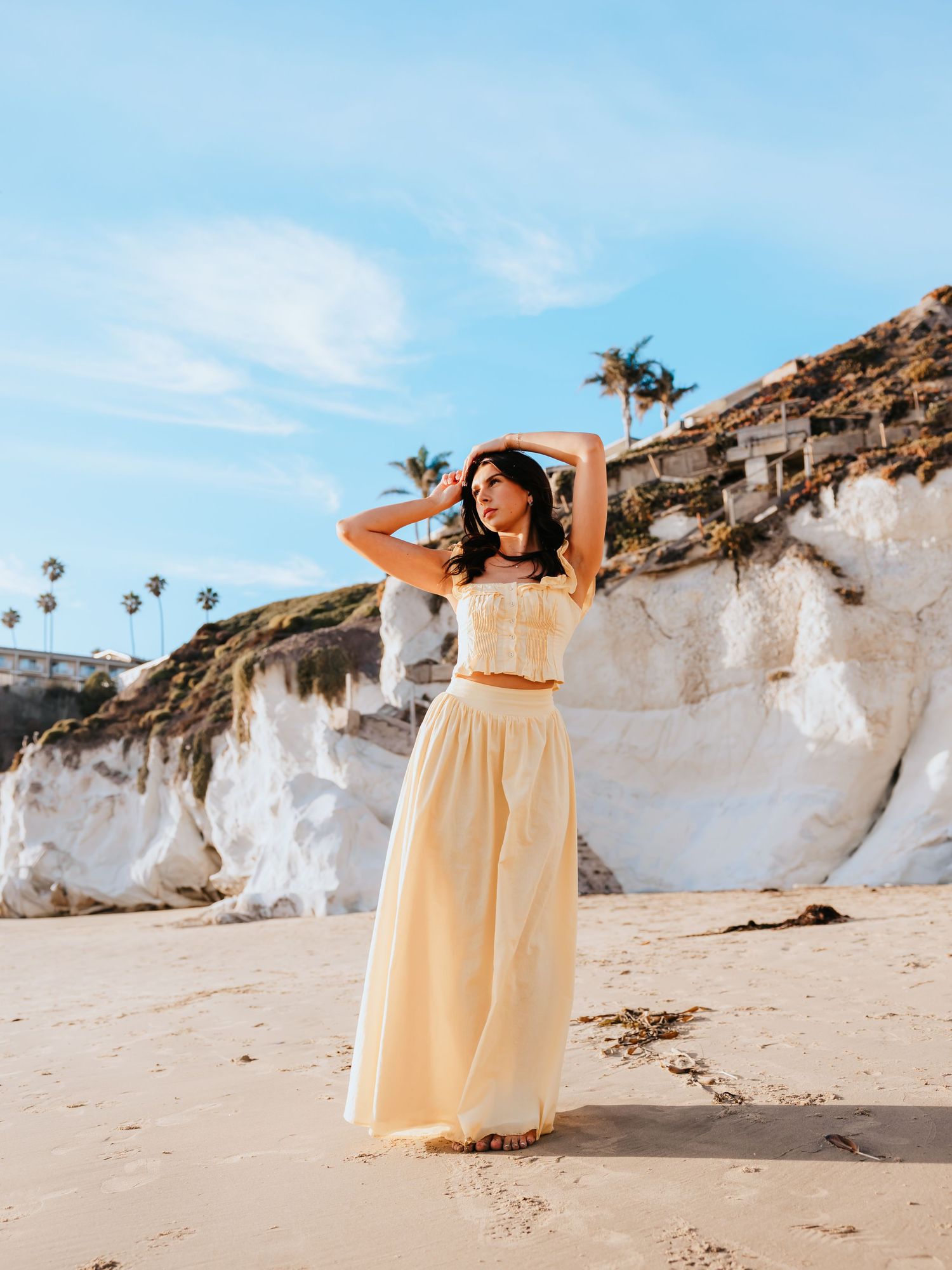 Woman in yellow crop top and maxi skirt posing on sandy beach with white cliffs and palm trees under blue sky.