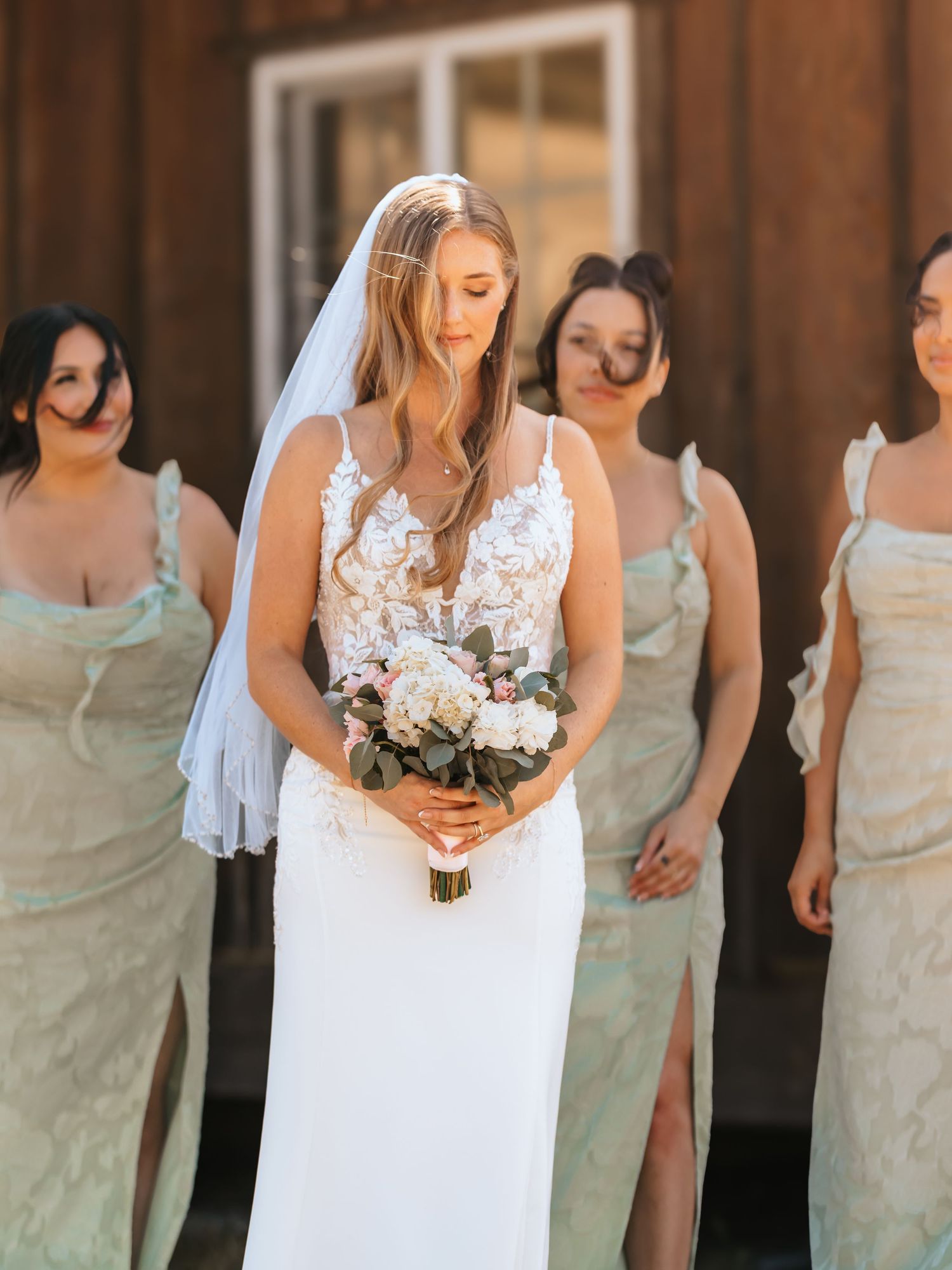 Bride in lace wedding gown holding floral bouquet standing before bridesmaids in sage green dresses.