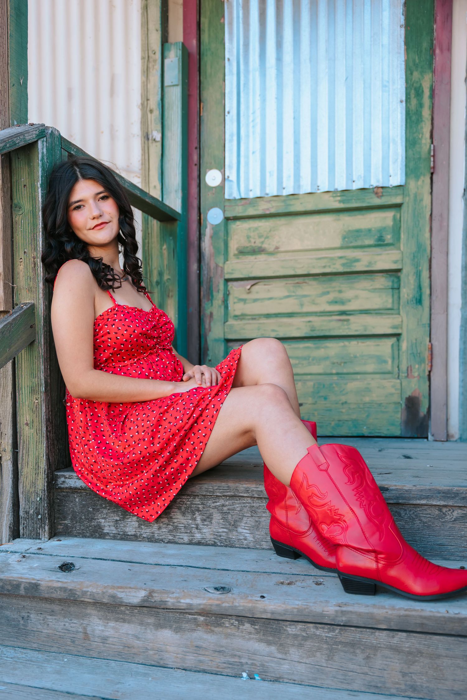 A young woman in a red dress and red cowboy boots sits on wooden steps of a rustic building.