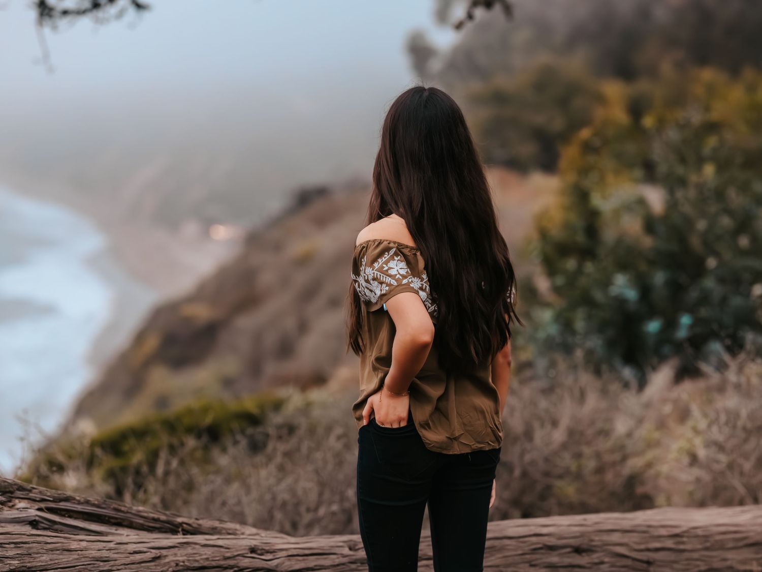 Woman with long dark hair standing on a cliff overlooking a scenic coastal landscape on a foggy day.