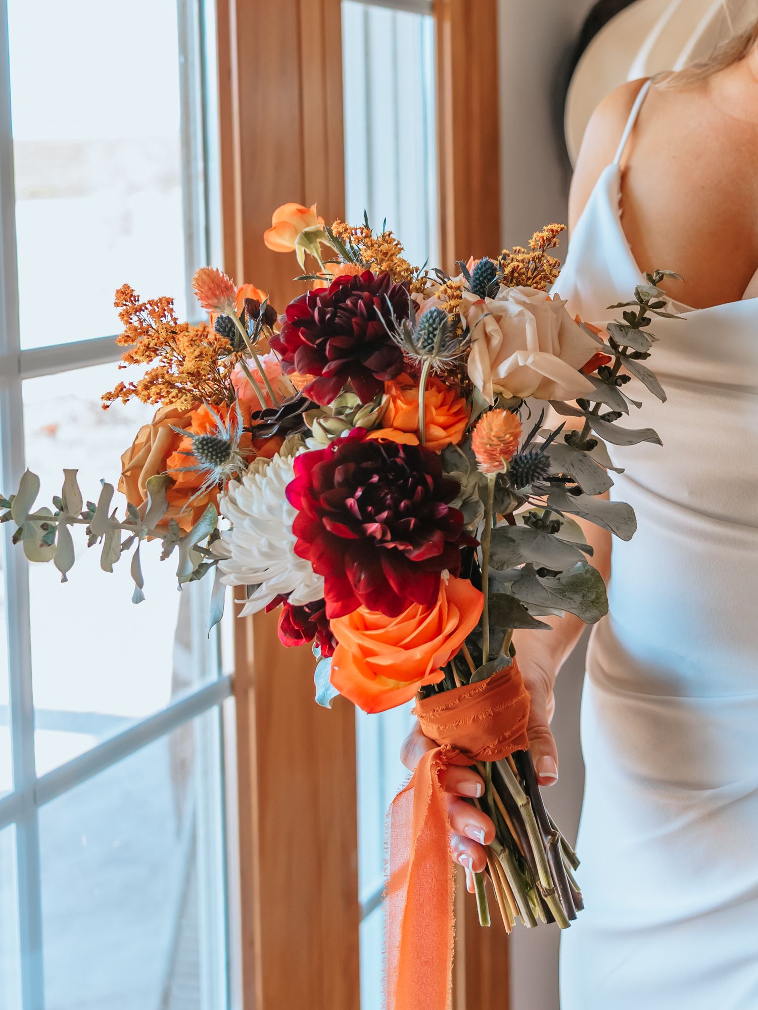 Bride holding a vibrant fall wedding bouquet with burgundy dahlias, orange roses, and eucalyptus near a window.