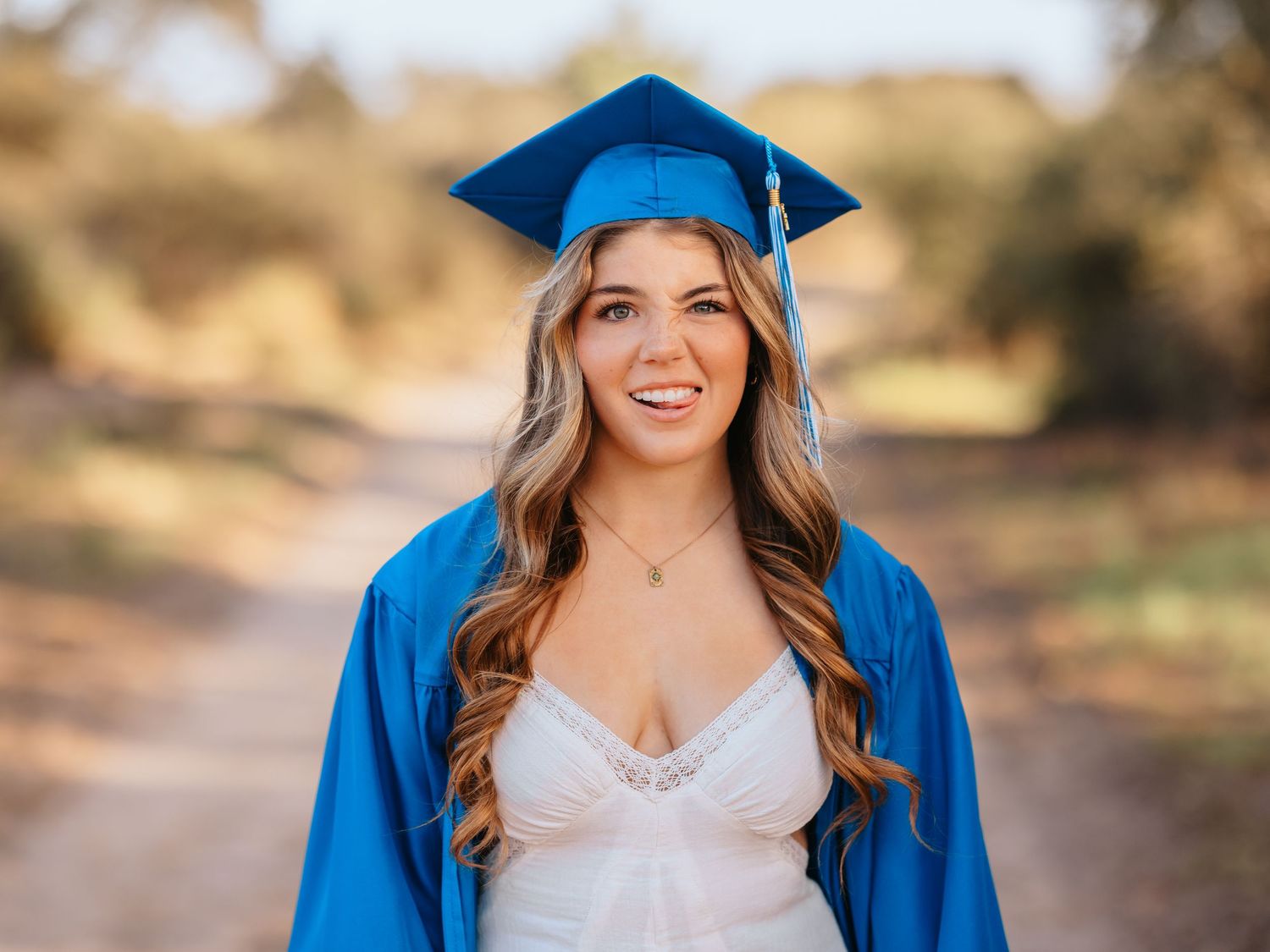 A graduate in bright blue cap and gown poses outdoors on a sunny day with trees in the background.