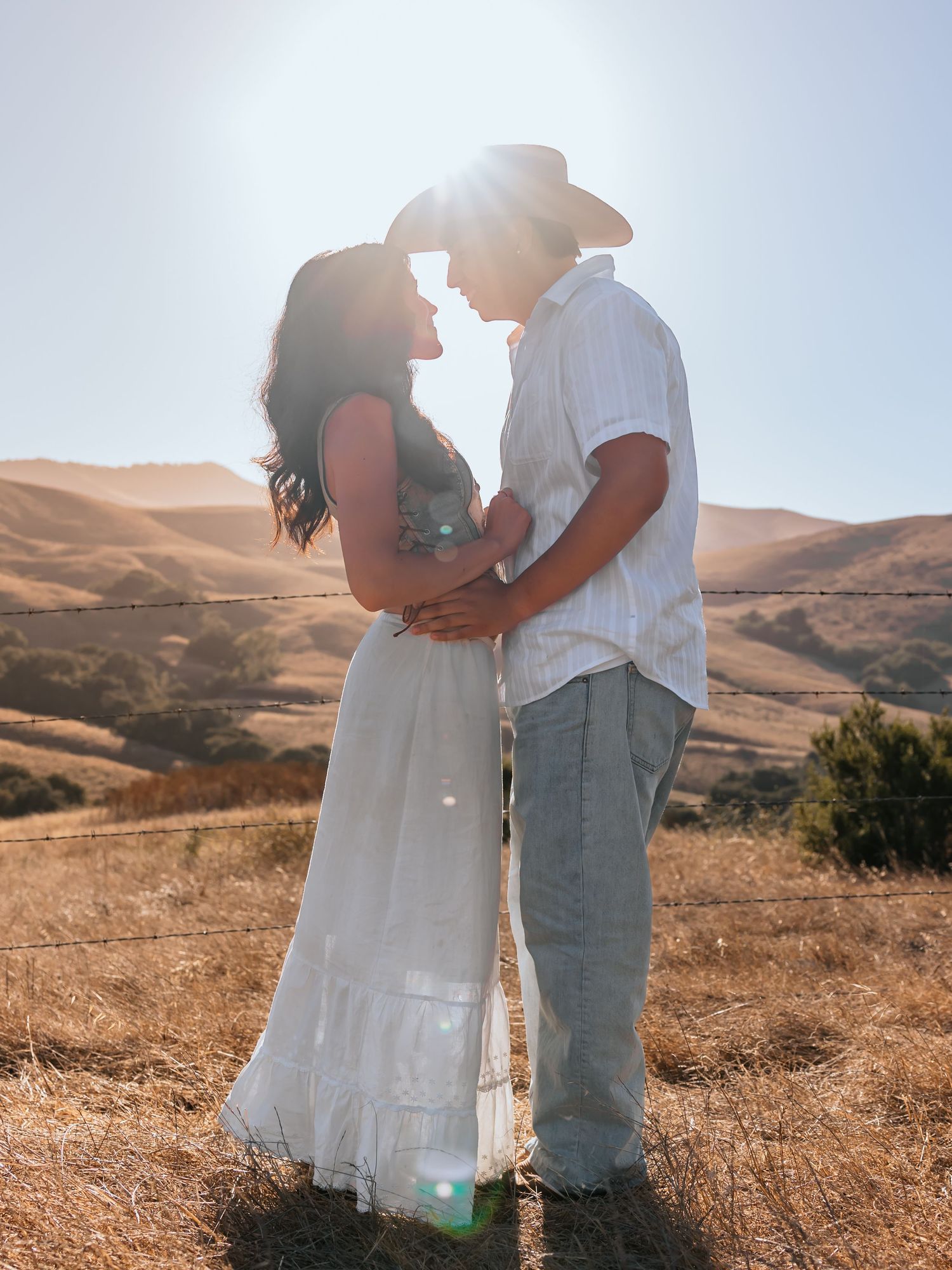 Couple in white embracing at golden hour with rolling hills backdrop, woman in maxi dress, man in cowboy hat.