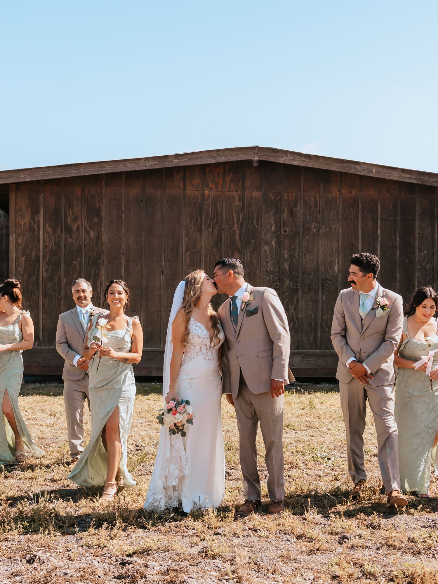 Bride and groom kissing while wedding party stands beside rustic barn under clear blue sky.