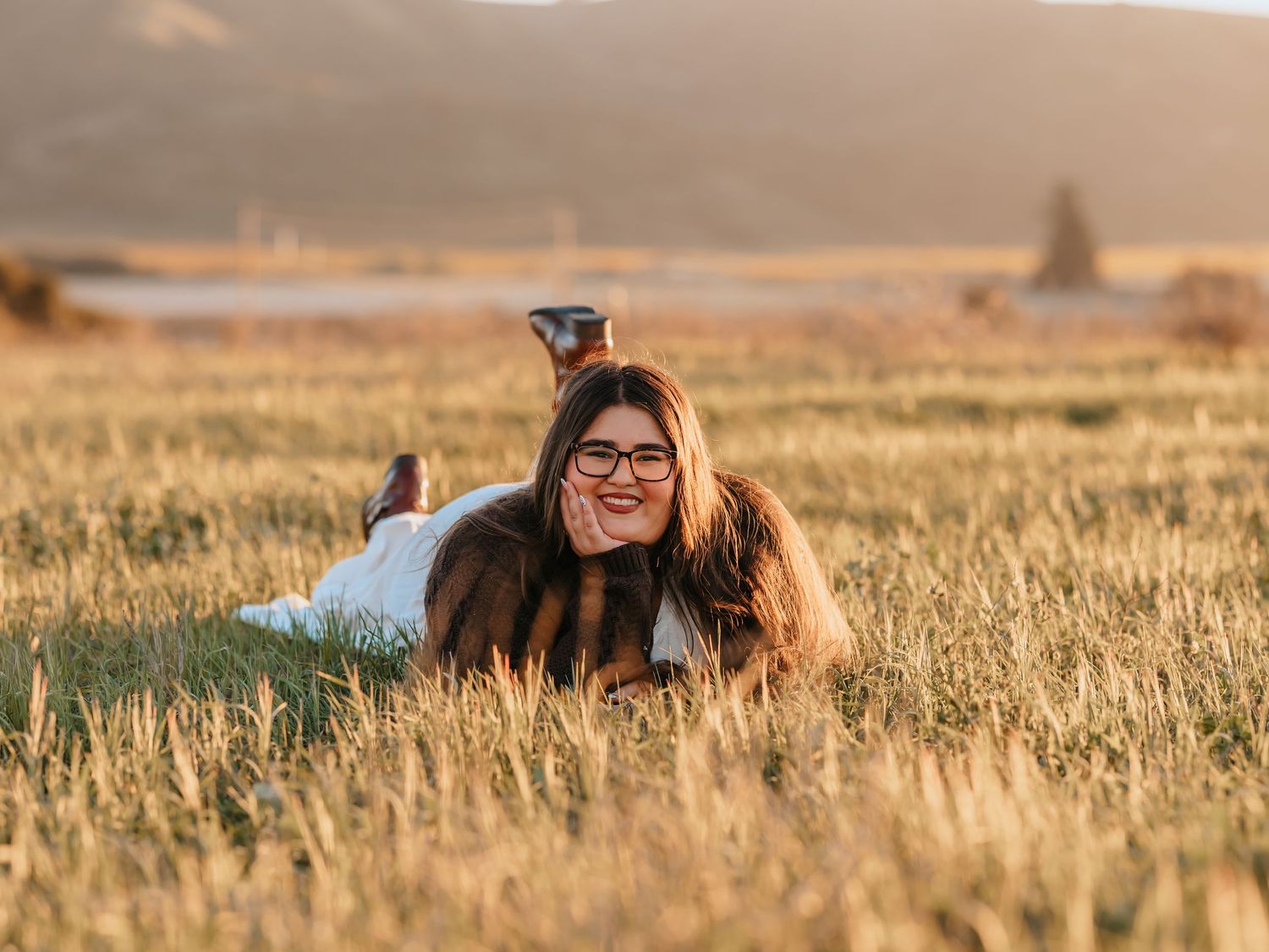 Young woman with glasses smiling while lying in golden field at sunset with mountains in background.