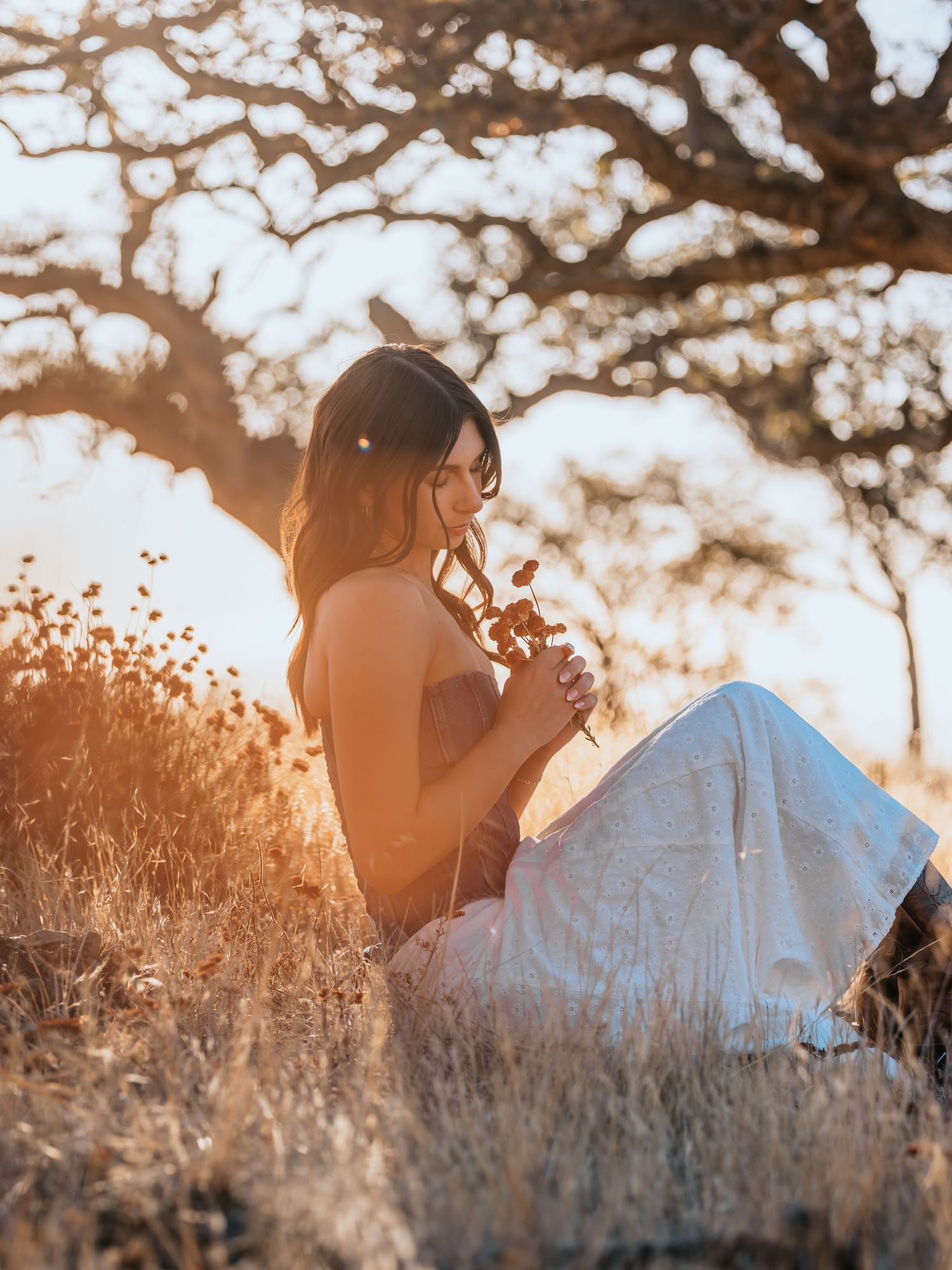 Woman in white pants sitting in golden field holding wildflowers at sunset beneath a tree.